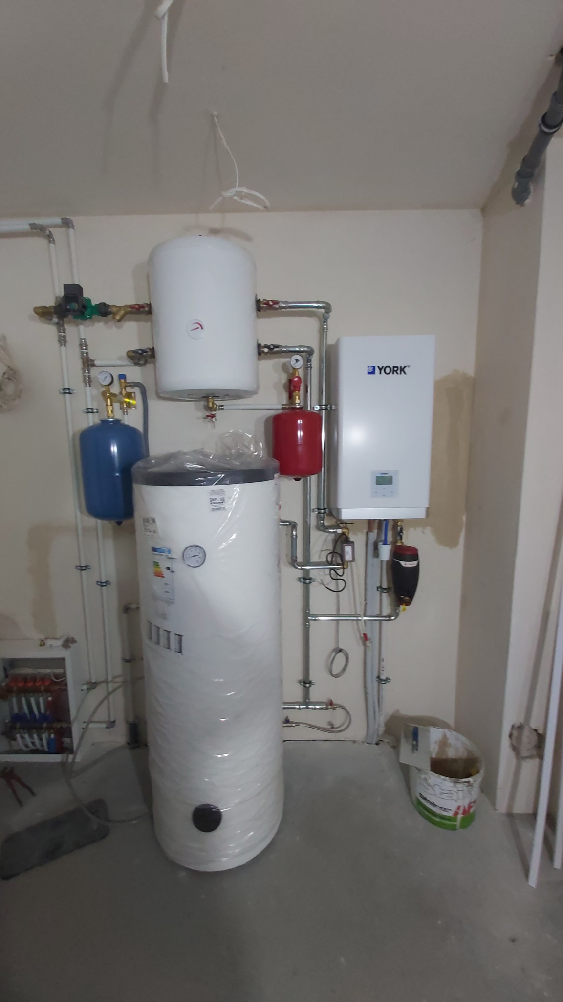 Złożona instalacja grzewcza z piecem York, zbiornikami (niebieski, czerwony, białe) i rurami. Nowa instalacja w pomieszczeniu z surowym wykończeniem.