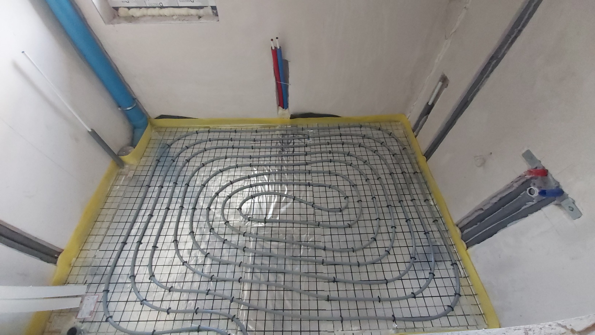 Szare rury ogrzewania podłogowego ułożone na czarnej siatce, zabezpieczone żółtą folią, widoczne pionowe rury instalacji na ścianie w surowym stanie.