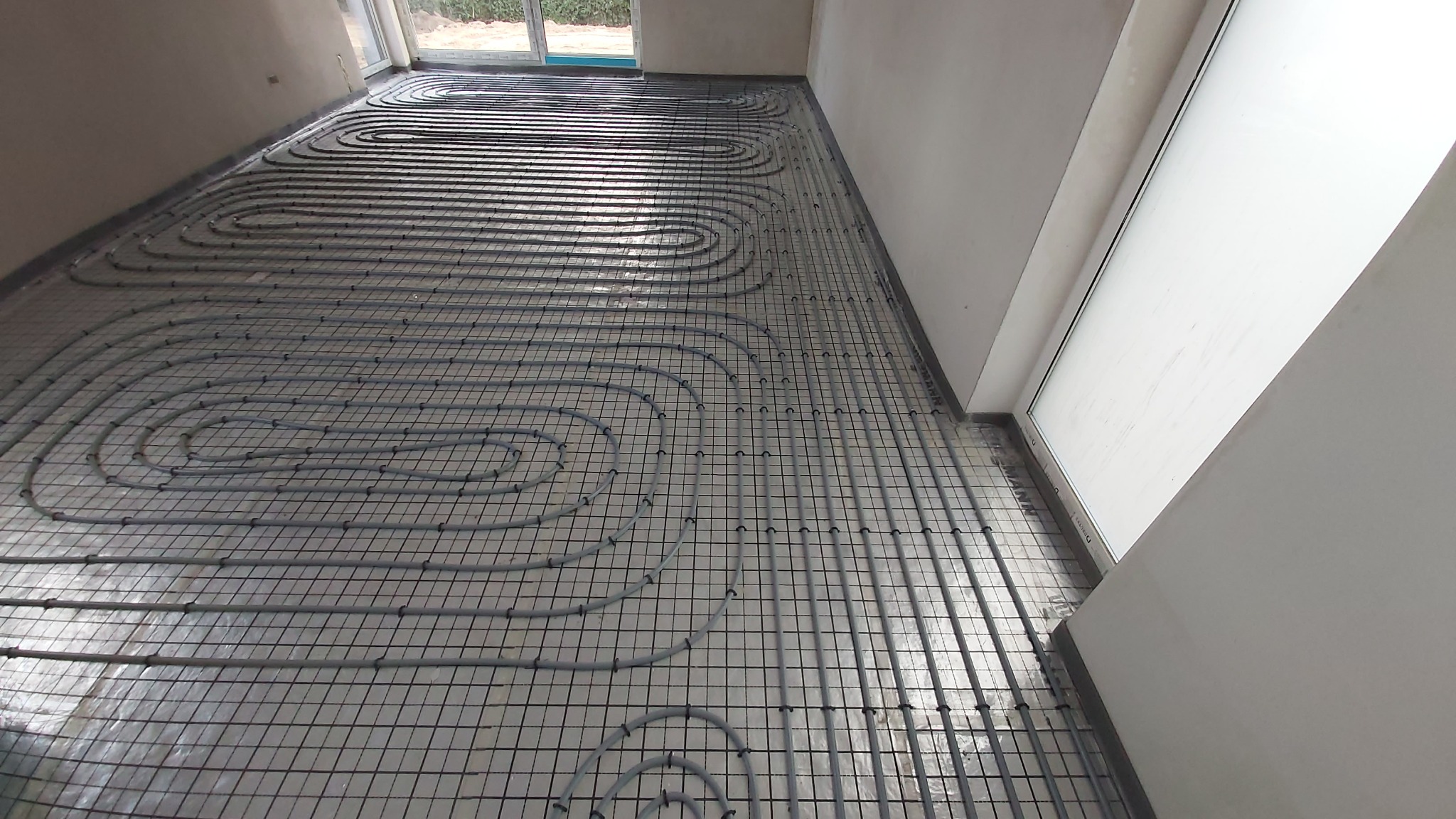 Szare rury ogrzewania podłogowego ułożone na srebrnej folii, przymocowane do czarnej siatki w jasnym pomieszczeniu z oknem. Widok z góry, ukośny kadr.
