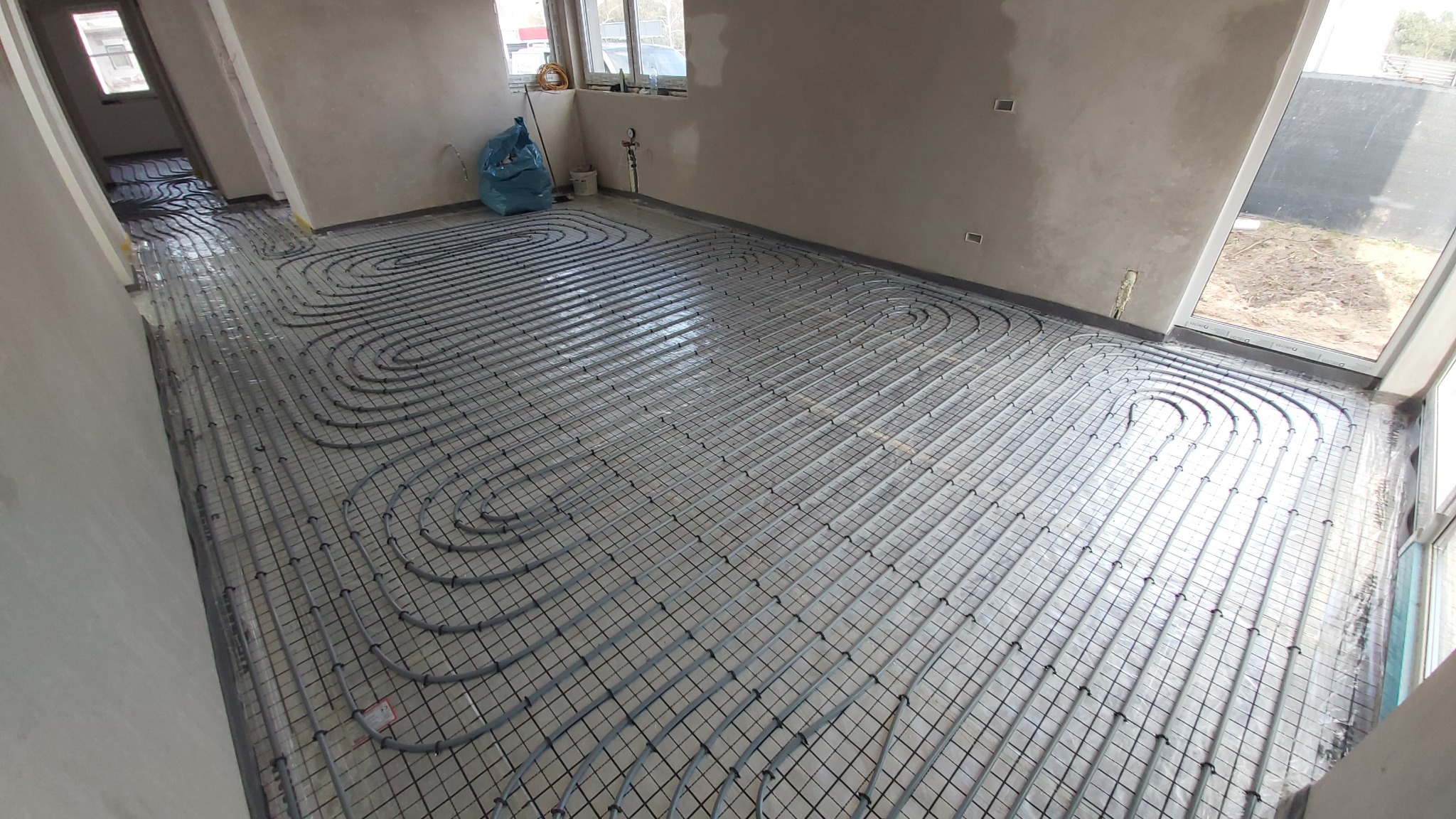 Szare rury ogrzewania podłogowego ułożone w pętle na siatce w surowym wnętrzu. Widoczne okna i worki budowlane. Etap instalacji przed wylewką.
