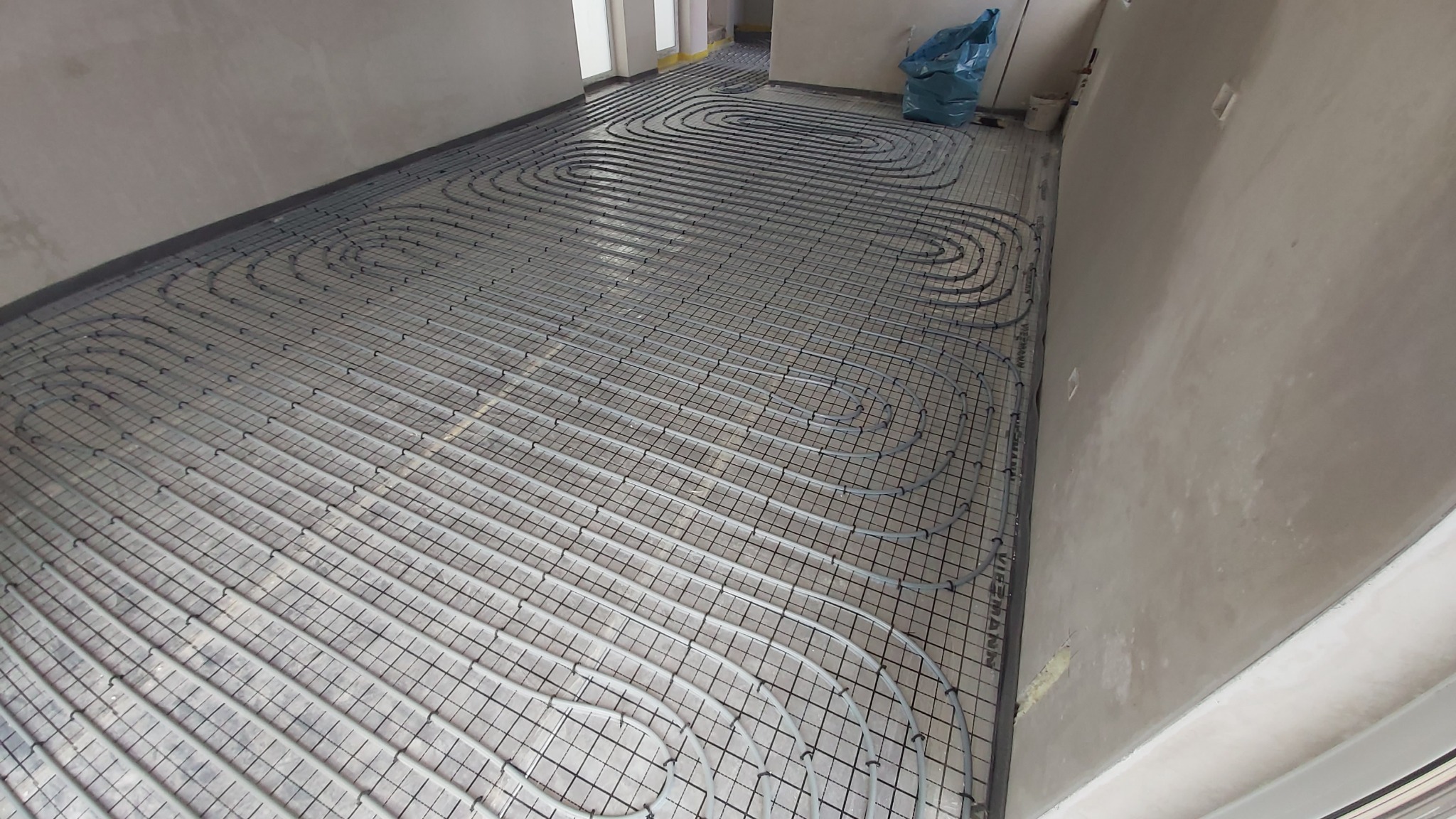 Szare rury ogrzewania podłogowego ułożone w pętle na metalowej siatce w surowym wnętrzu. Widoczne okna i worki z materiałami budowlanymi w tle.