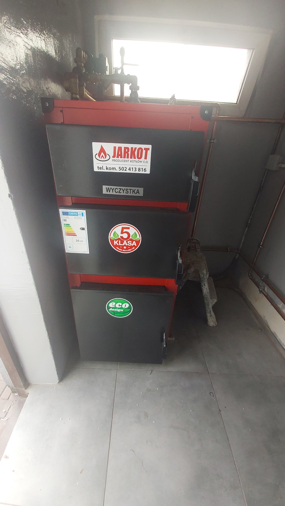 Czerwono-czarny piec CO marki Jarkot z naklejką '5 Klasa' i 'eco design' w kotłowni. Widoczne rury i okno. Piec z bliska, z widocznymi detalami.