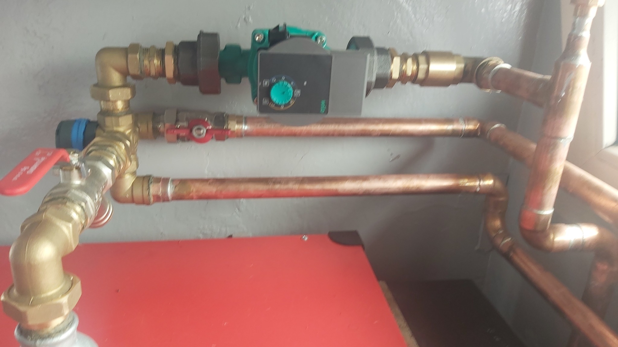 Miedziane rury instalacji grzewczej z zaworami i pompą obiegową, zamontowane na szarej ścianie nad czerwonym zbiornikiem. Widoczne złącza i elementy regulacyjne.