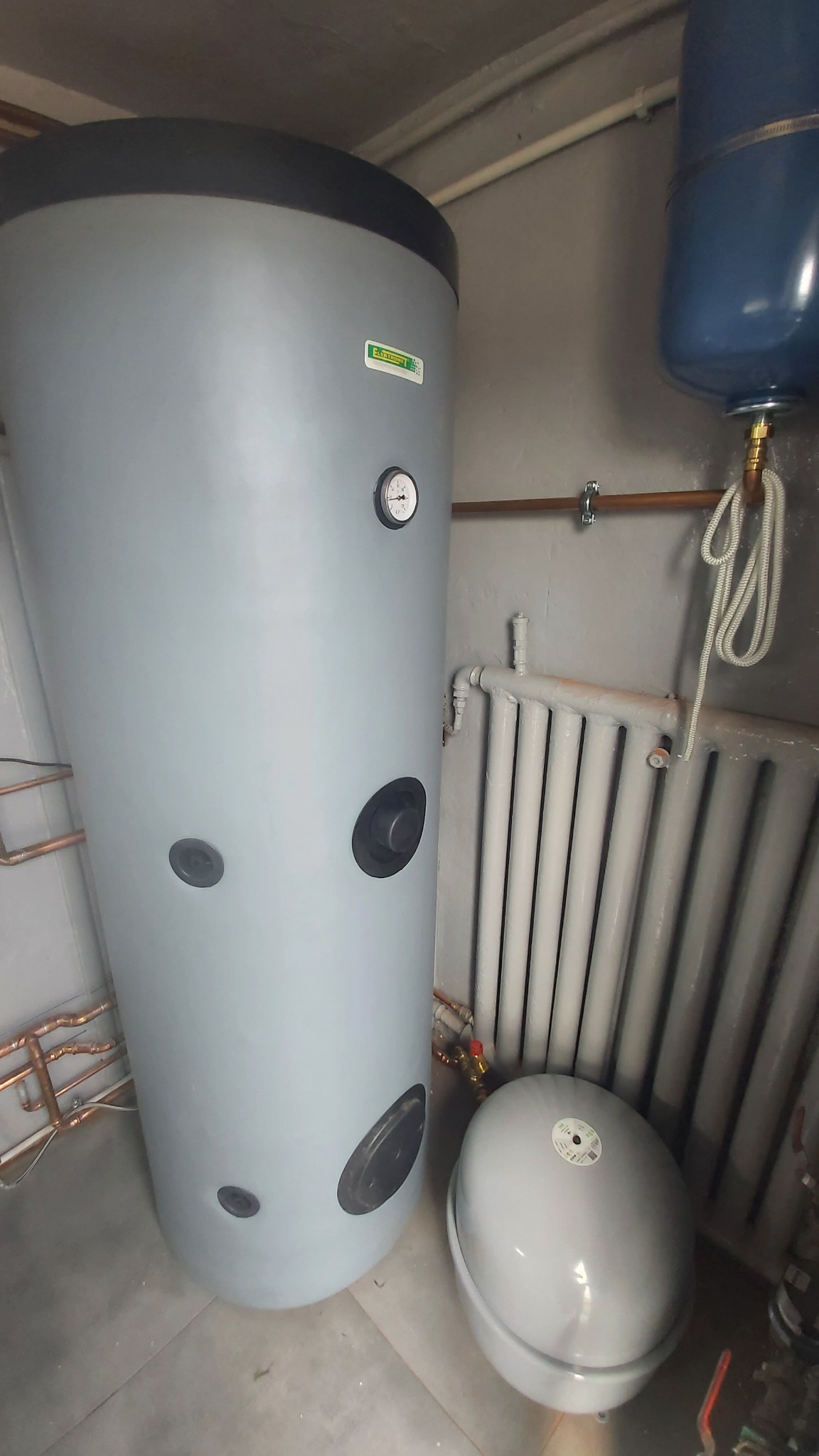 Szary, cylindryczny zasobnik ciepłej wody użytkowej z manometrem, obok grzejnik żeberkowy i zbiornik wyrównawczy w kotłowni. Widoczne rury miedziane.