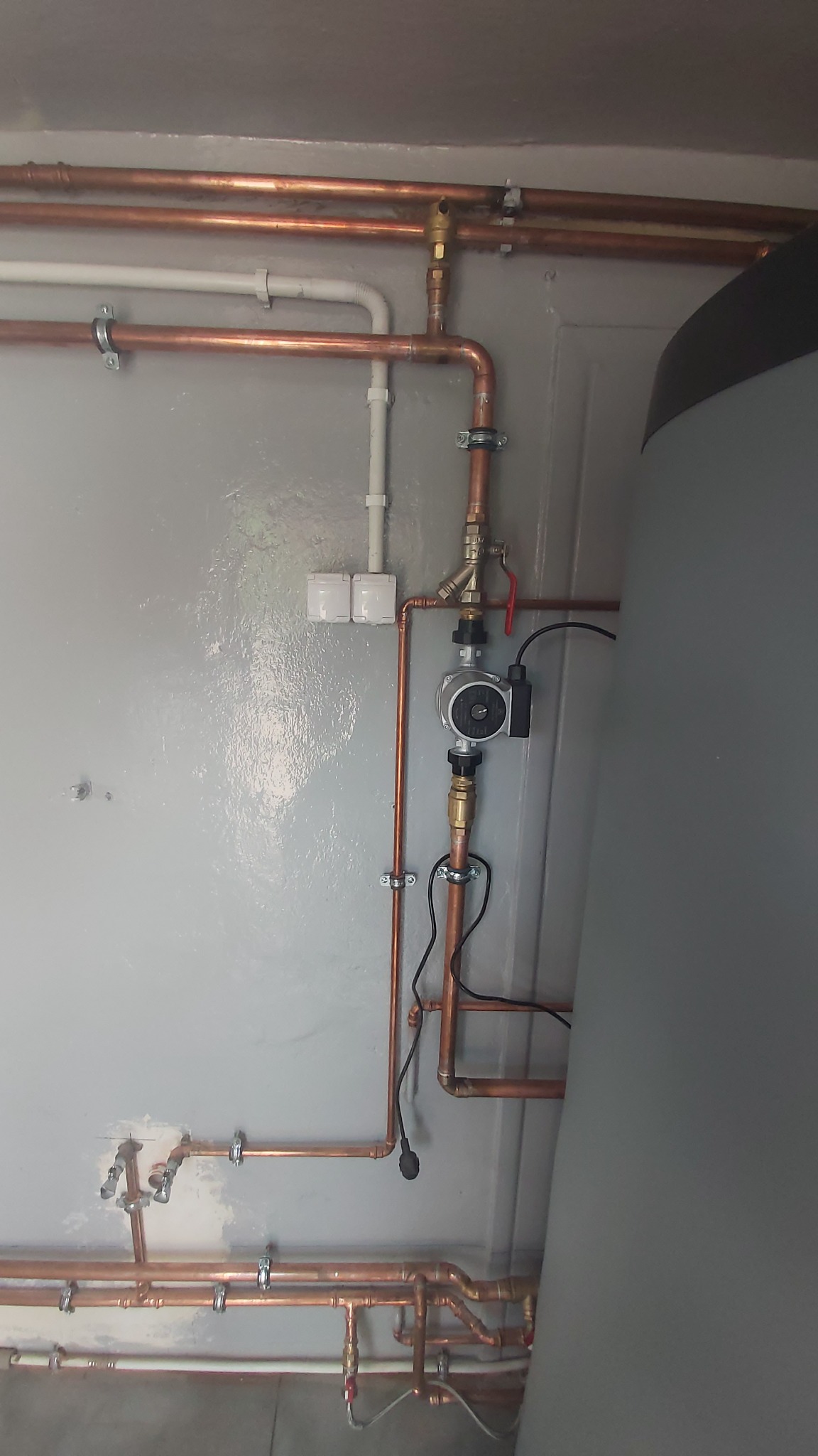 Miedziane rury instalacji CO z pompą obiegową i zaworami, zamocowane na szarej ścianie. Widoczny fragment zbiornika akumulacyjnego.