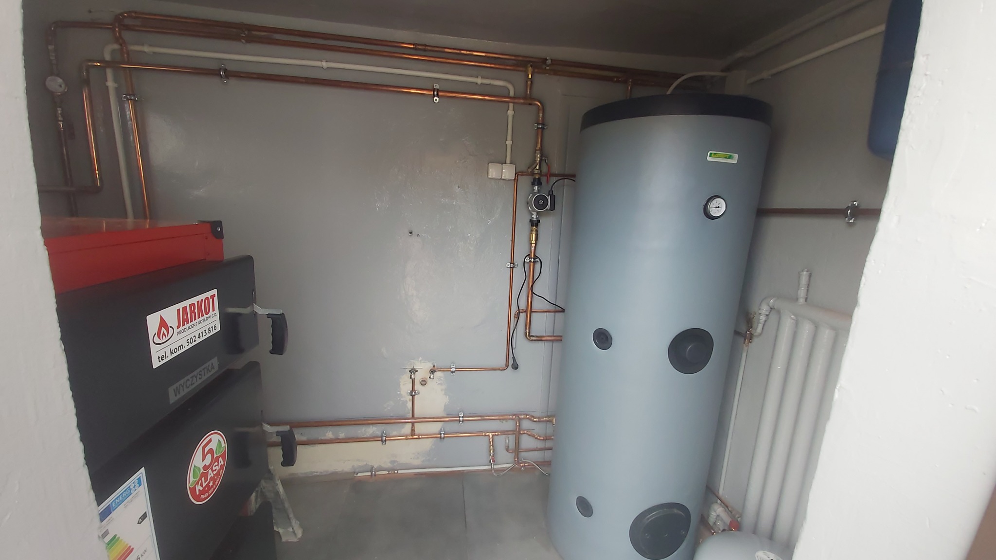 Kotłownia z piecem Jarkot, zasobnikiem ciepłej wody i miedzianą instalacją grzewczą. Widoczne rury, zawory i manometr. Szare ściany i biały grzejnik.