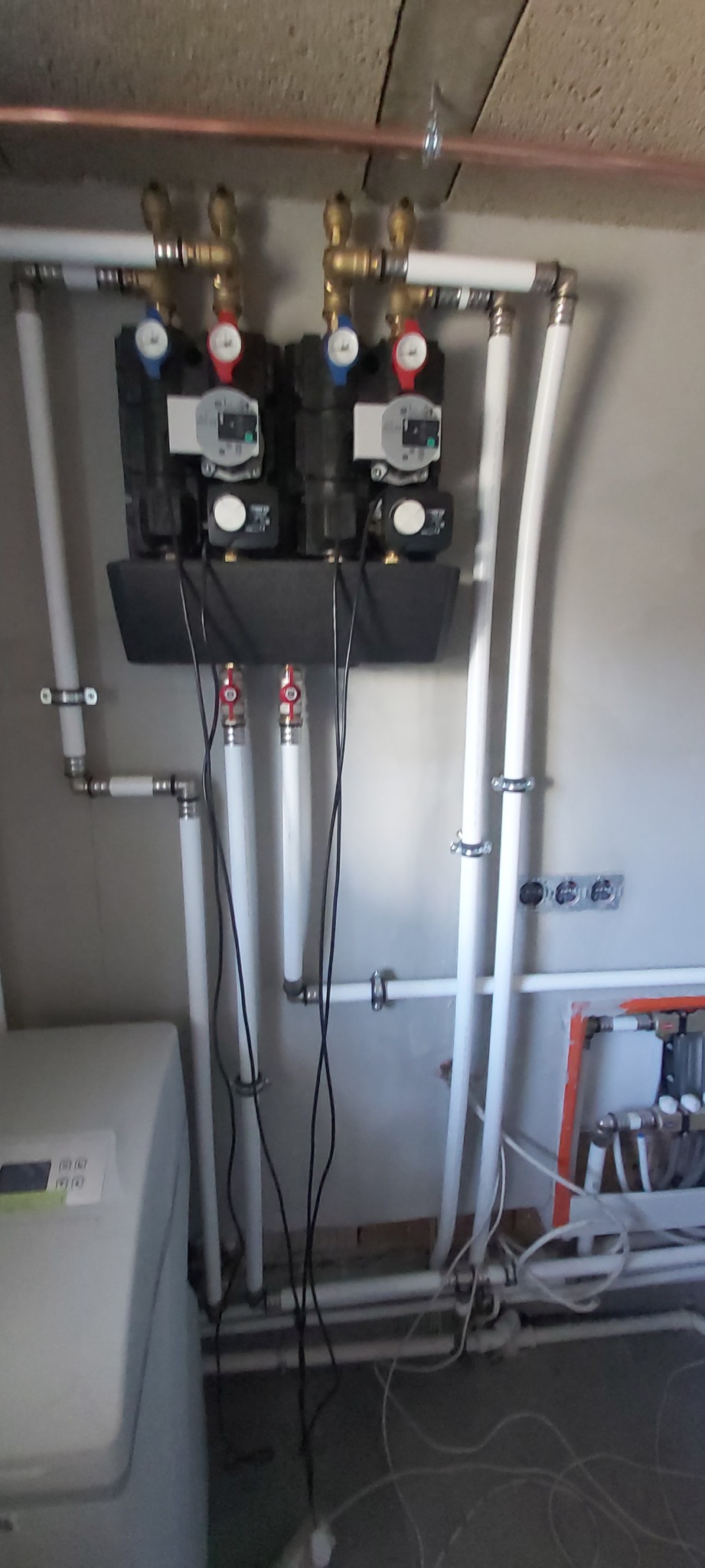 Rozdzielacz ogrzewania podłogowego z dwoma pompami obiegowymi, rurami z tworzywa i miedzianymi elementami, zamontowany na szarej ścianie w kotłowni.