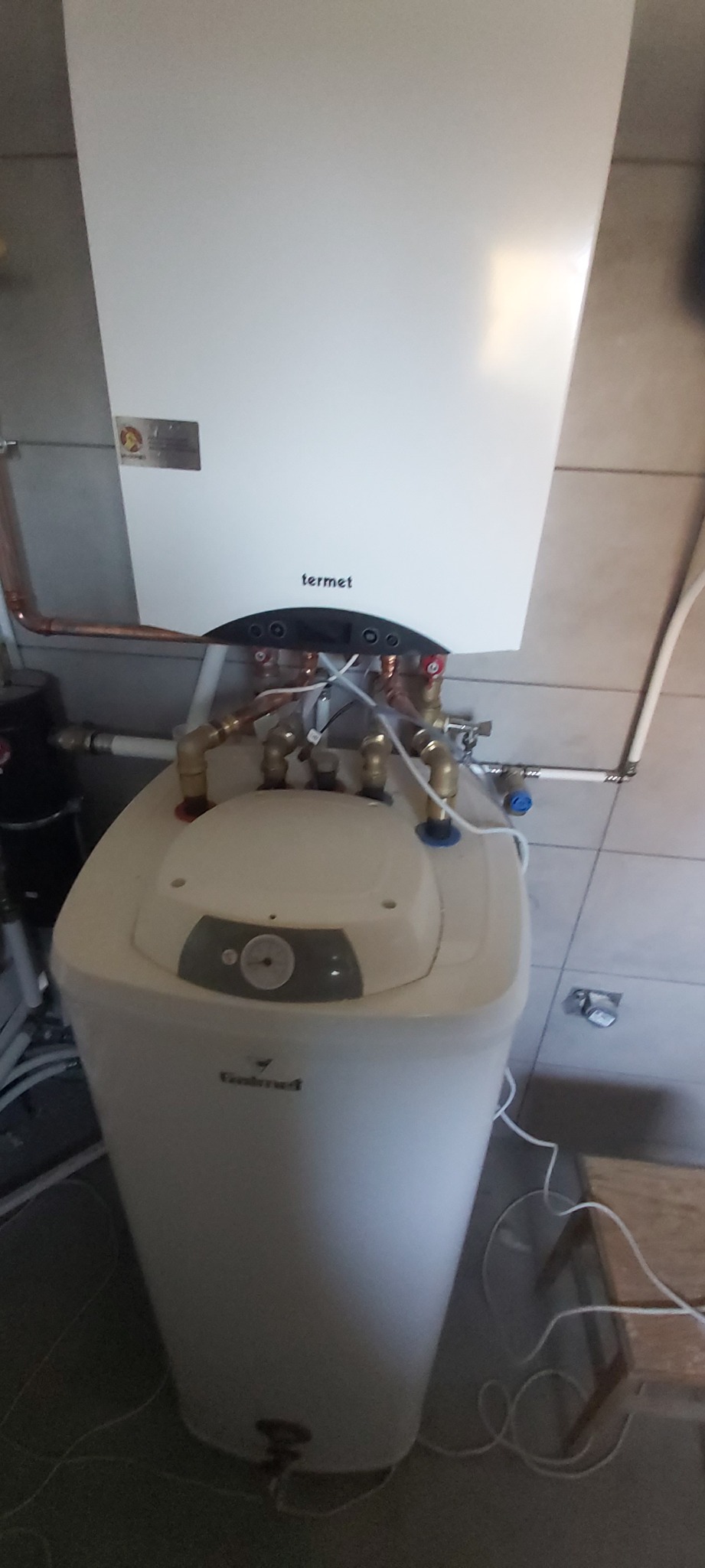 Biały piec gazowy Termet zamontowany na ścianie z szarymi płytkami, z widocznymi miedzianymi rurami i elementami instalacji. U dołu biały zasobnik wody.