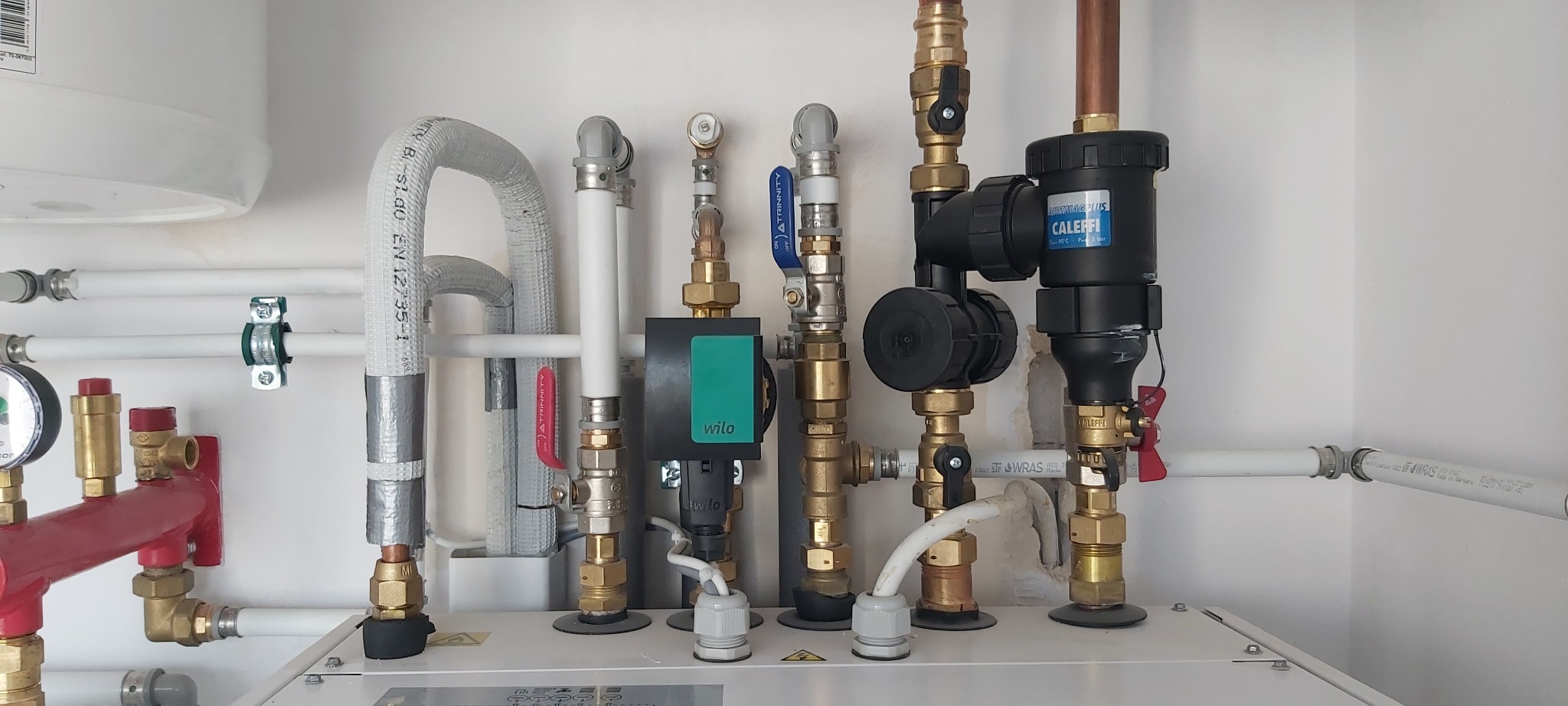 Złożona instalacja grzewcza z rurami miedzianymi i plastikowymi, zaworami, pompą obiegową Wilo i filtrem Caleffi, zamontowana na białej ścianie. Widoczne różne elementy instalacji.