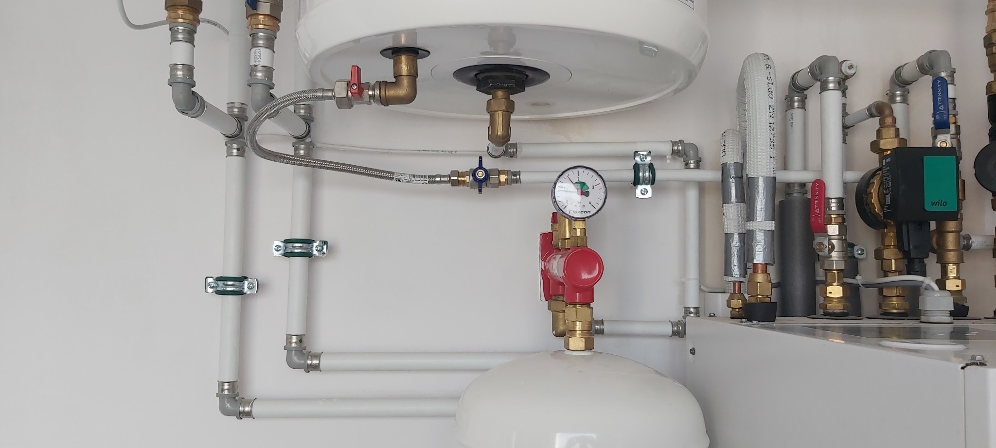Białe rury instalacji CO/CWU z zaworami, manometrem i pompą obiegową, zamontowane na białej ścianie. Widoczne elementy kotła i zasobnika.