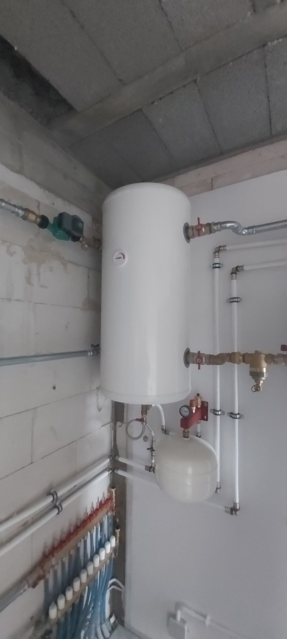 Biały bojler z podłączonymi rurami miedzianymi i plastikowymi, zamontowany na ścianie z białych bloczków. Widoczny także rozdzielacz ogrzewania podłogowego.