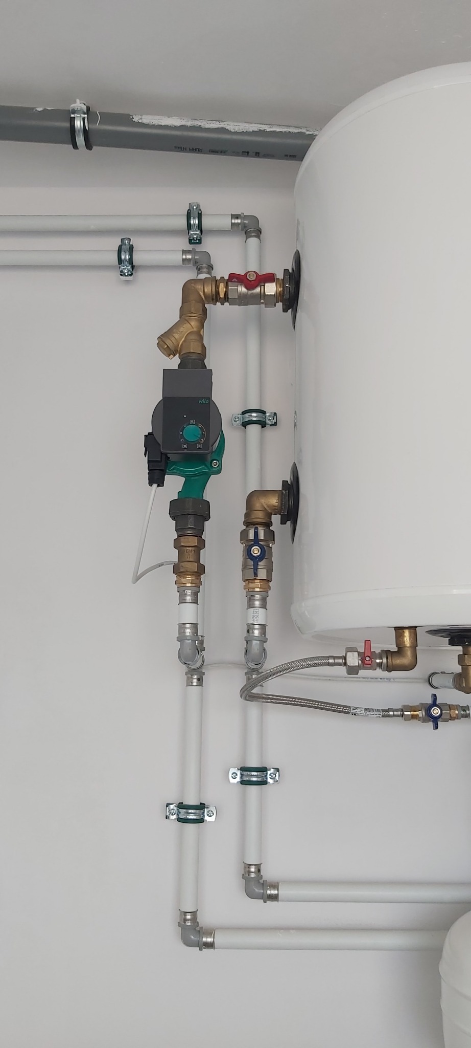 Biały bojler z podłączonymi rurami i zaworami, widoczny zielony obiegowy pompa Wilo. Instalacja hydrauliczna w jasnym pomieszczeniu, rury mocowane klamrami do ściany.
