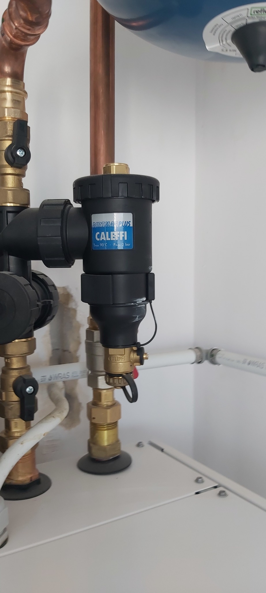 Zamontowany filtr Caleffi Dirtmag Plus na miedzianych rurach instalacji grzewczej, widoczne zawory i białe rury PCV. Filtr zamontowany na białej ścianie.