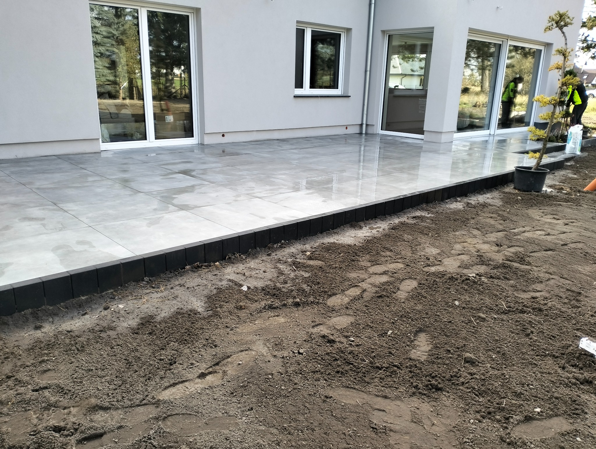 Nowoczesny taras z szarych płyt, obrzeża z ciemnej kostki. Częściowo przygotowany teren pod dalsze prace. Duże okna przesuwne w tle.