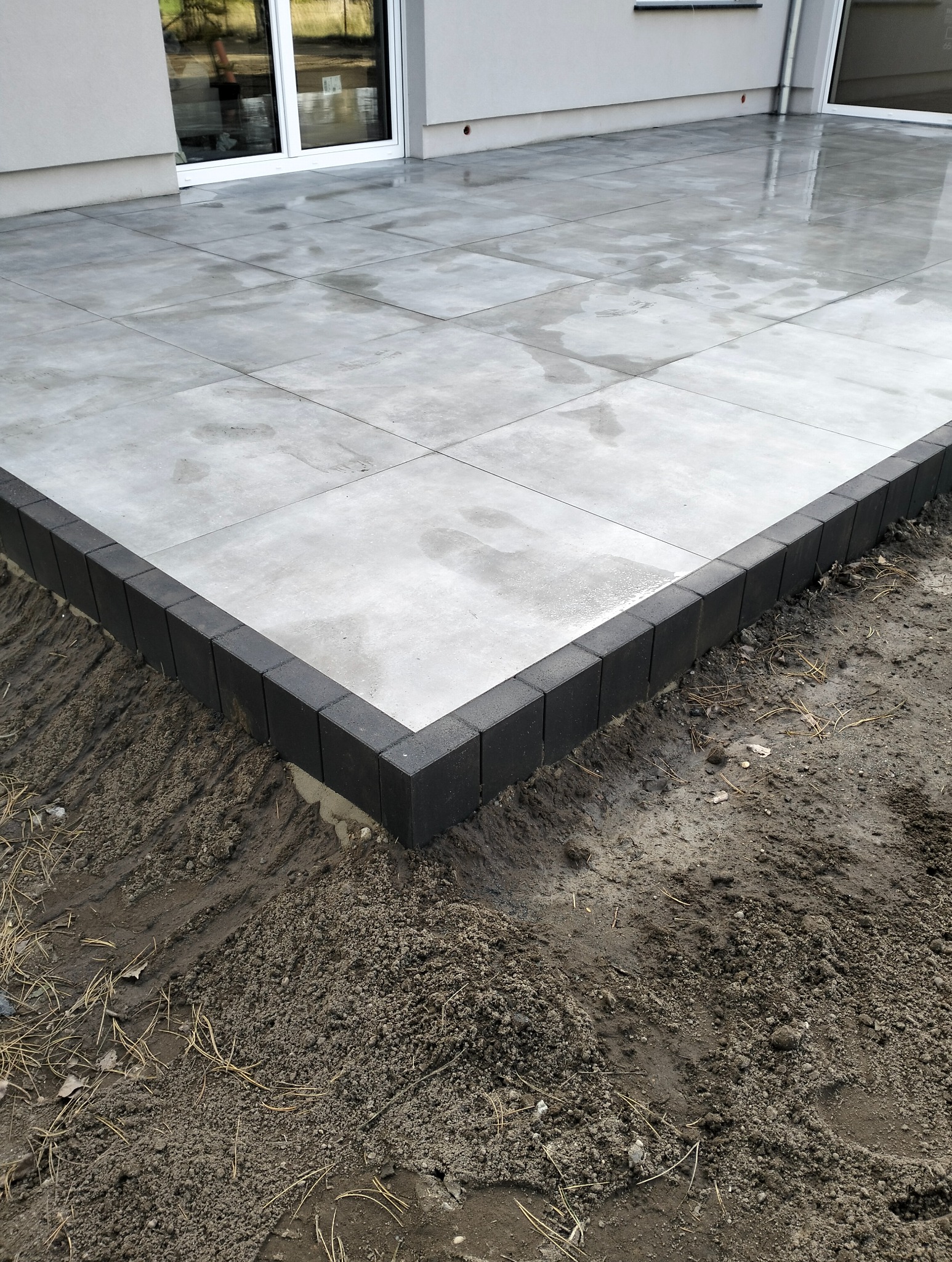 Nowoczesny taras z szarych płyt betonowych obramowany czarną kostką, widok na świeżo obsypaną ziemię. Minimalistyczny design, surowy klimat.