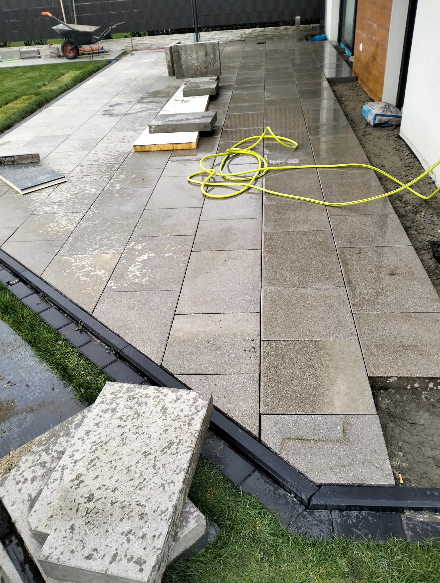 Szare, prostokątne płyty chodnikowe w trakcie układania, z widocznym systemem drenażowym i żółtym wężem ogrodowym. Część płyt jeszcze nieułożona.