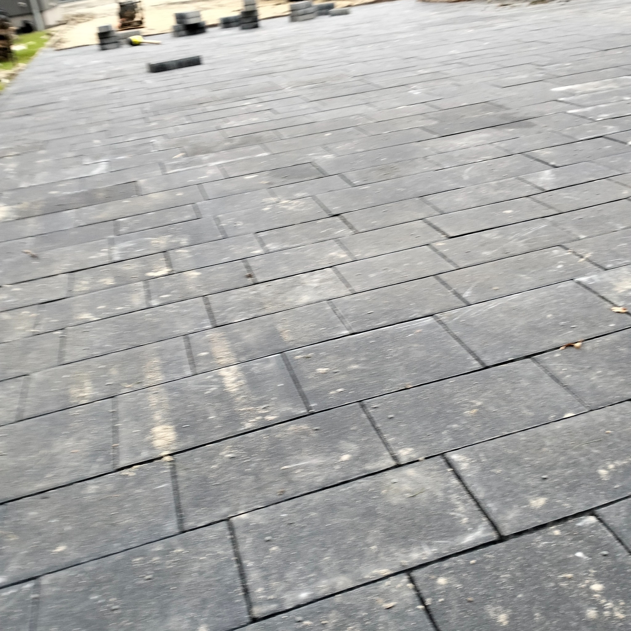 Ułożona, szara kostka brukowa w regularnym wzorze, z widocznymi fugami. W tle stos kostki i narzędzia, sugerujące trwające prace wykończeniowe.