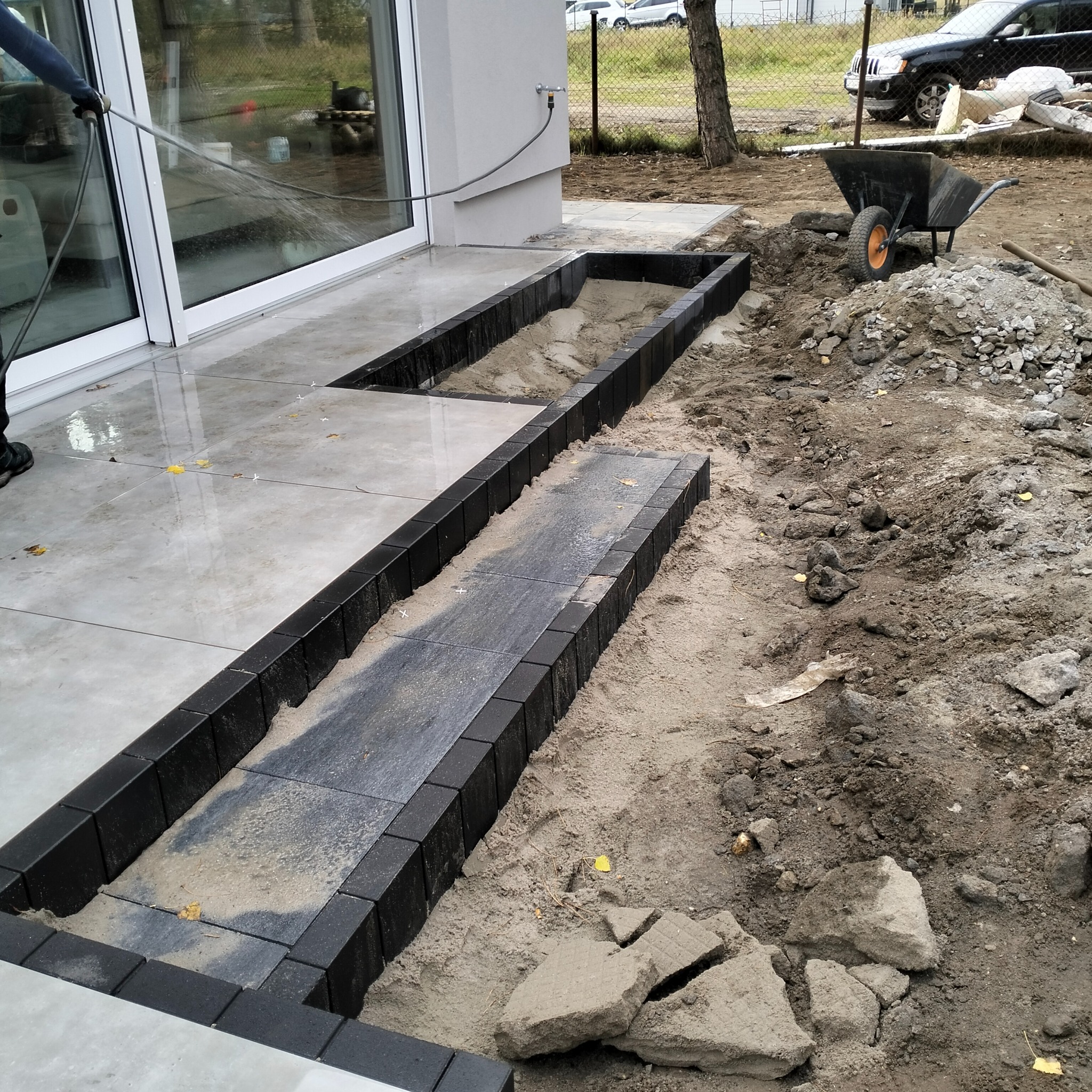 Układanie kostki brukowej: czarne obrzeża i szare płyty na piasku, widoczne narzędzia i wąż z wodą. Etap prac brukarskich przy budynku.