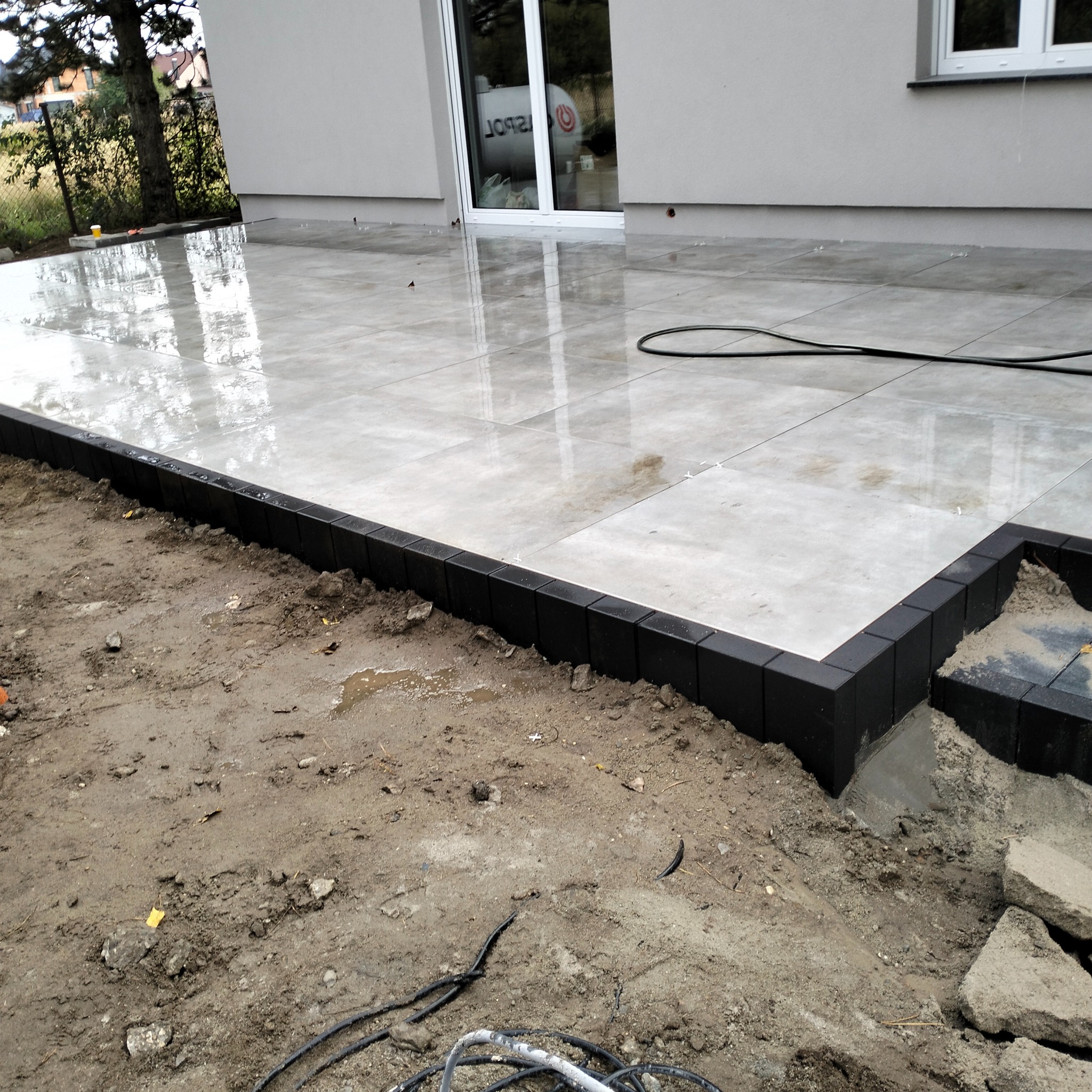 Wykończony taras z jasnych, kwadratowych płyt, obramowany czarną kostką. Widoczny fragment ogrodu z ziemią i kablami. Świeżo ułożona nawierzchnia po deszczu.
