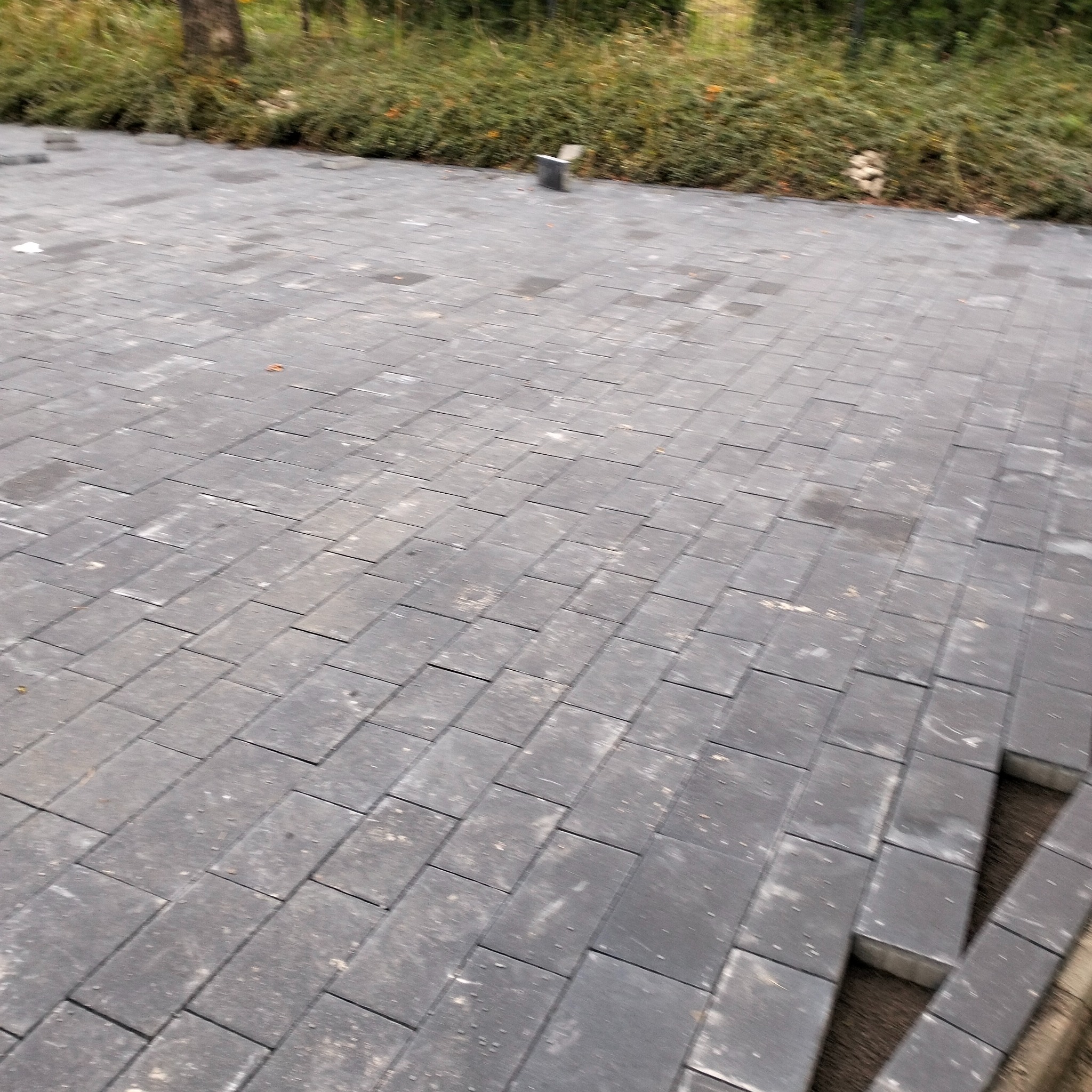 Ułożona, szara kostka brukowa w regularnym wzorze, z fragmentem niedokończonej krawędzi po prawej stronie. W tle widoczna zieleń krzewów i drzew.