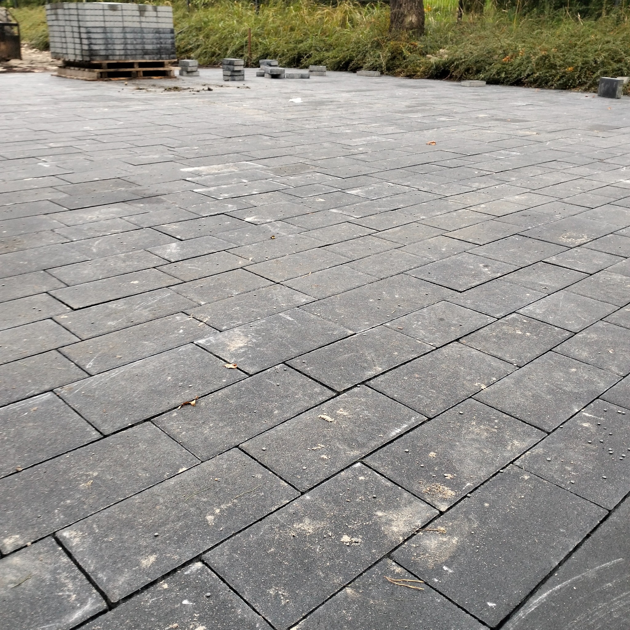 Ułożona, szara kostka brukowa na posesji, z paletą kostki w tle i zielenią. Widok z perspektywy, ukazujący regularny wzór i estetyczne wykończenie nawierzchni.