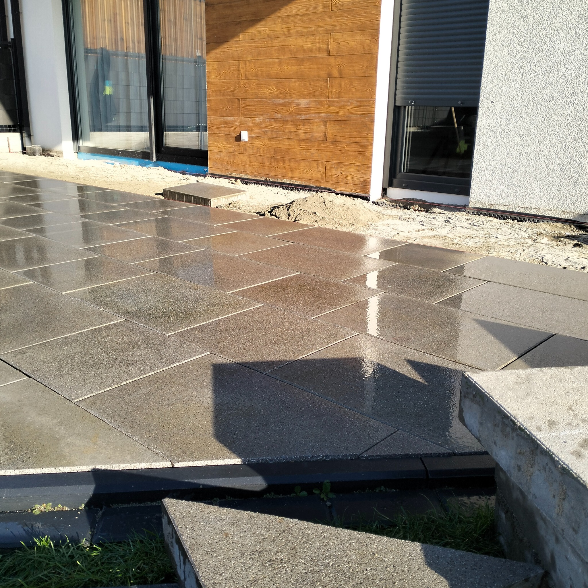 Świeżo ułożona, mokra kostka brukowa w odcieniach szarości, odbijająca światło słoneczne. Widoczne fragmenty domu z drewnianą elewacją i elementami wykończeniowymi.