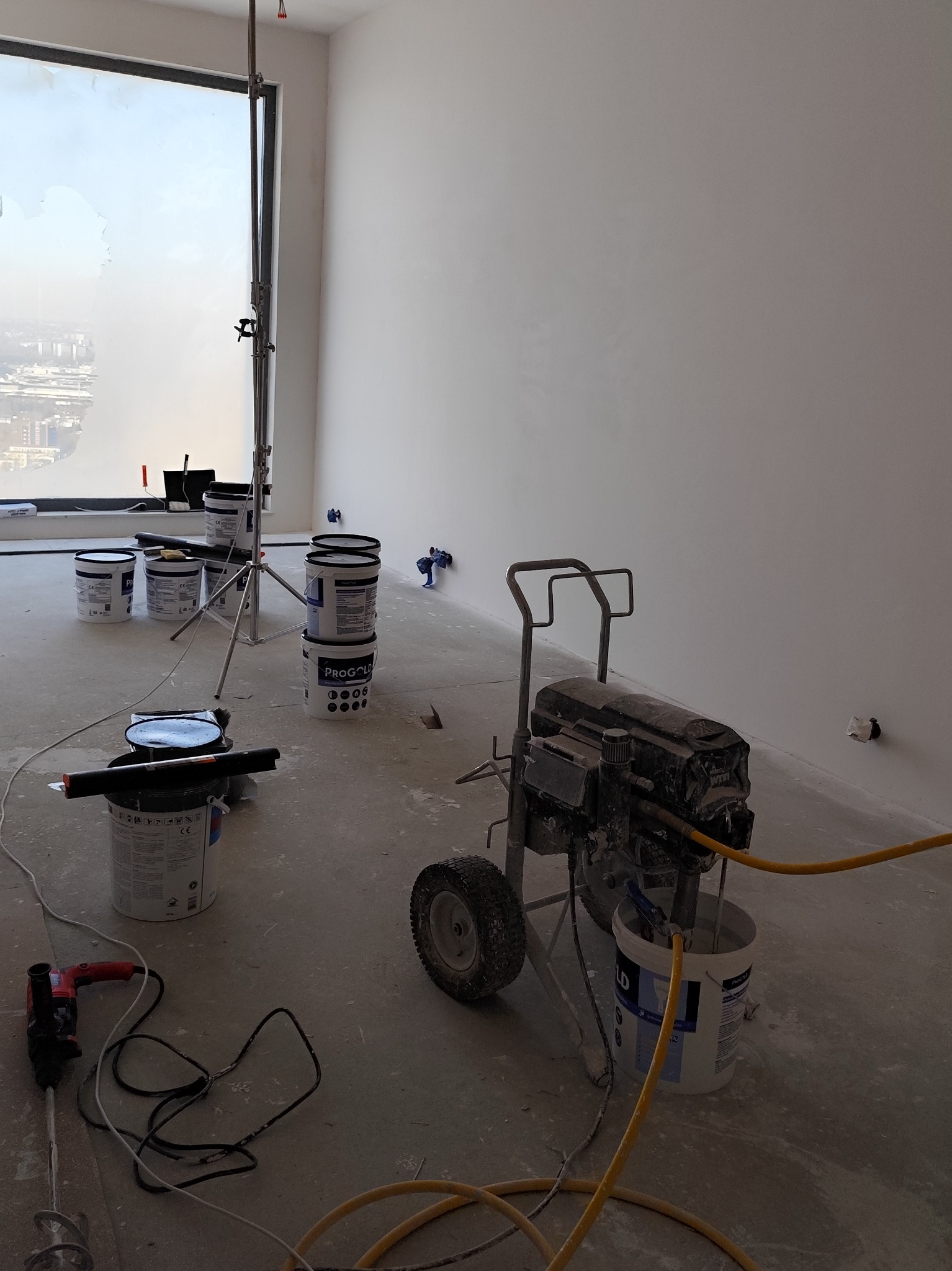 Białe ściany po malowaniu natryskowym w jasnym wnętrzu. Widoczny agregat malarski, puszki z farbą i narzędzia malarskie. Duże okno z widokiem na miasto.