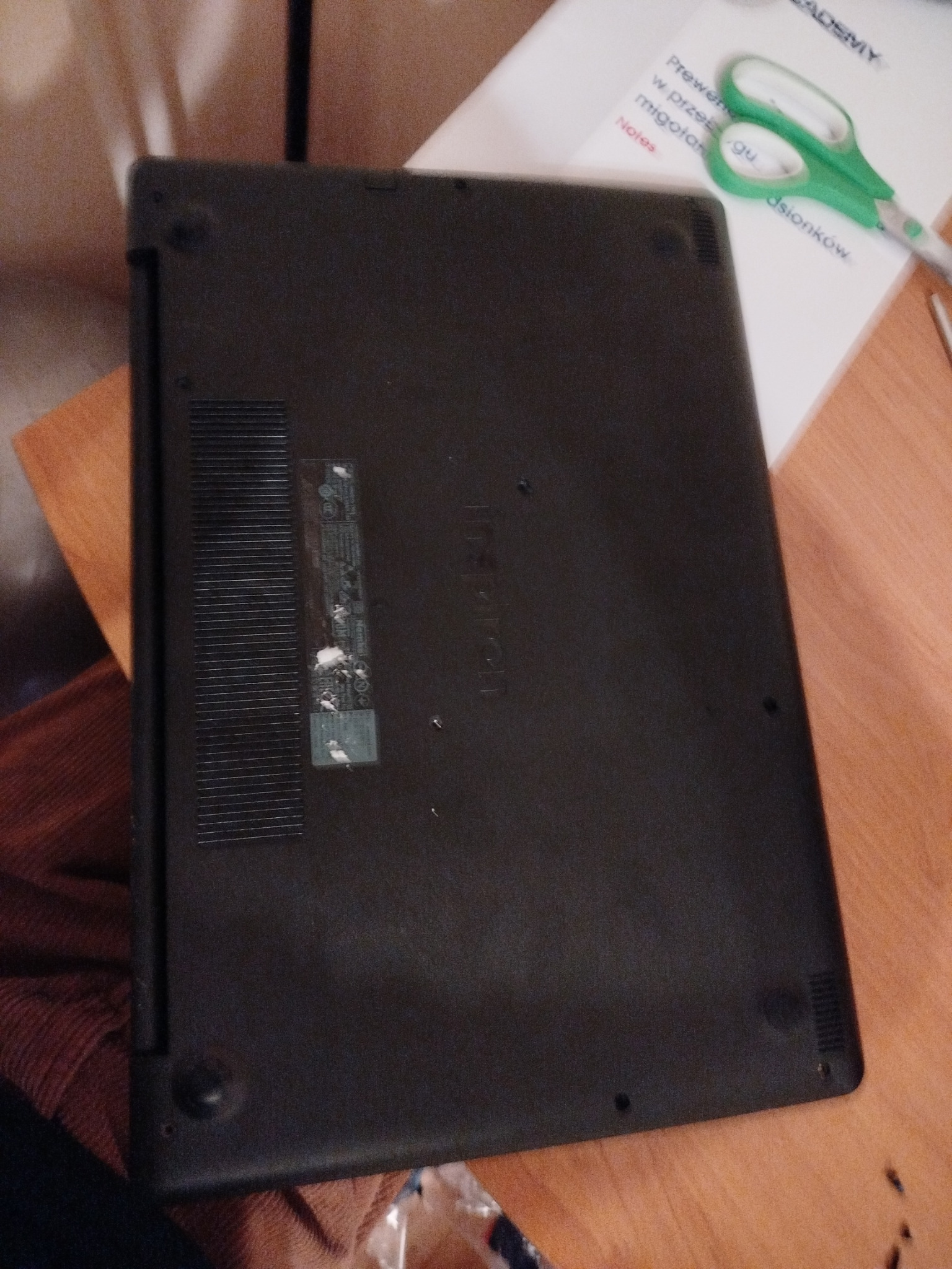 Spód czarnego laptopa marki Lenovo z widoczną naklejką i otworami wentylacyjnymi, leżącego na biurku obok nożyczek i notatek.