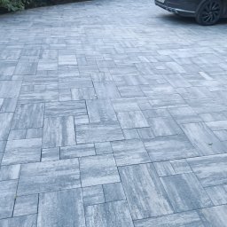 VOLODYMYR DIDUKH - Ułożona szara kostka brukowa na podjeździe, z zaparkowanym samochodem w tle i zielenią po bokach. Widok z perspektywy lekko z góry, ukazujący wzór ułożenia.