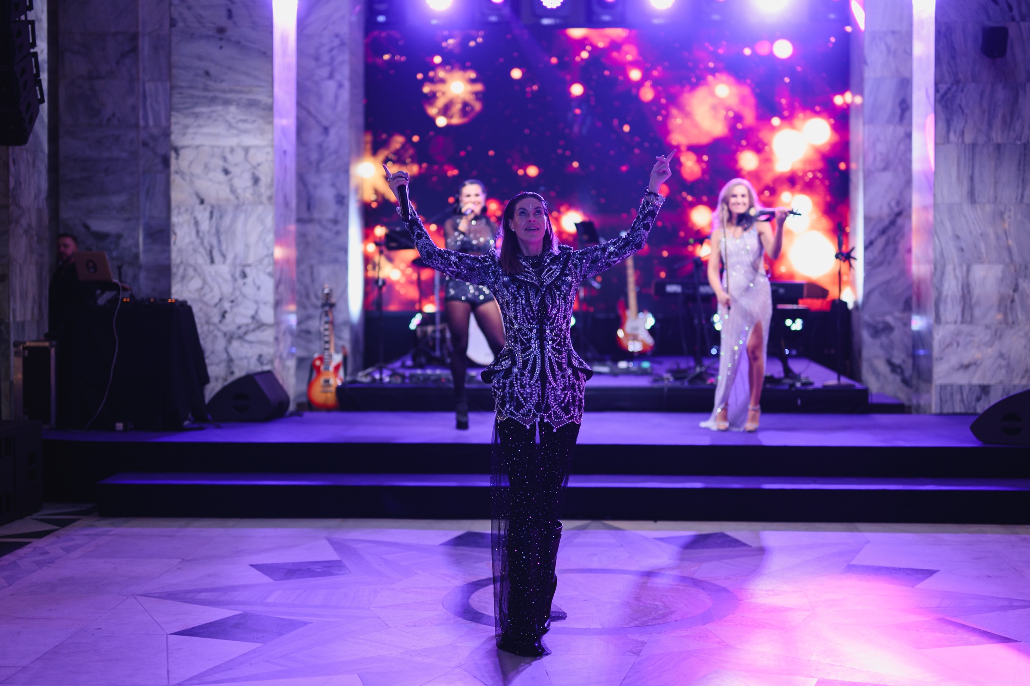 Zespół muzyczny na scenie podczas występu na imprezie. Wokalistka w błyszczącej kreacji, skrzypaczka i wokalistka w tle. Scena oświetlona fioletowymi światłami.