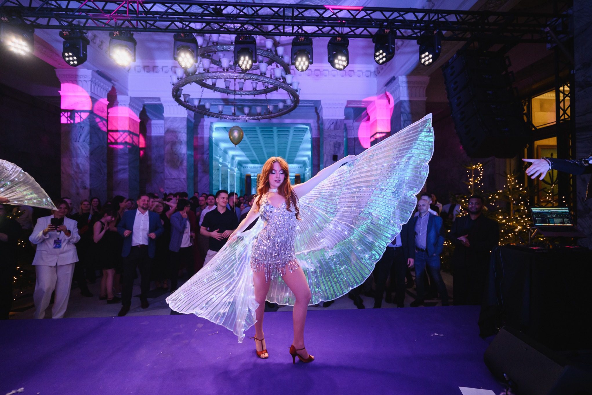 Kobieta w błyszczącej sukience i skrzydłach LED na scenie podczas eventu. W tle publiczność, oświetlenie sceniczne i dekoracje.