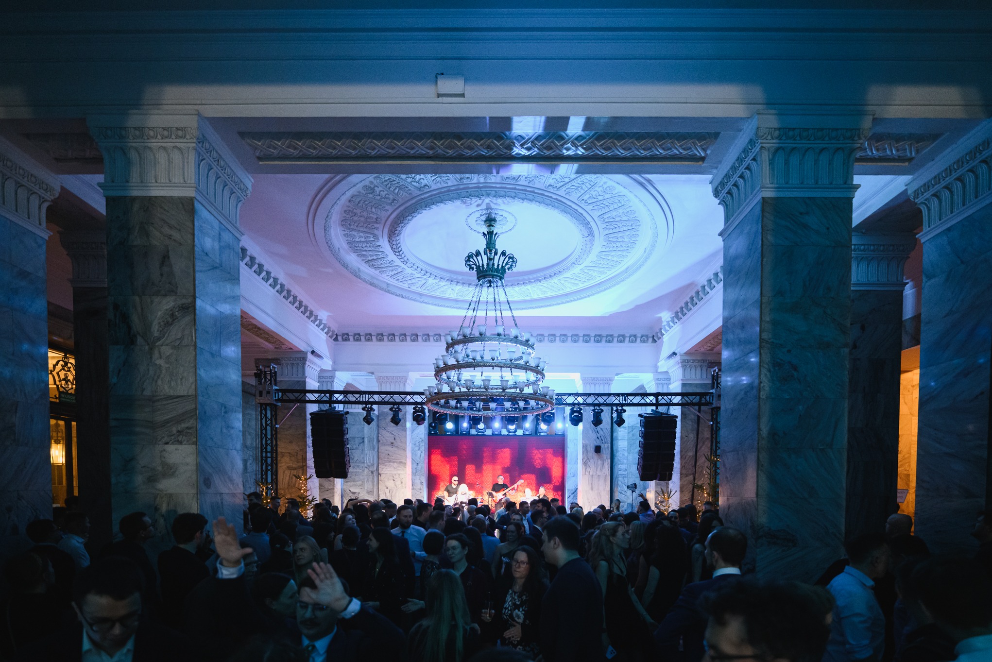 Imponująca sala balowa z marmurowymi kolumnami, kryształowym żyrandolem i sceną z zespołem muzycznym grającym dla zgromadzonego tłumu gości.