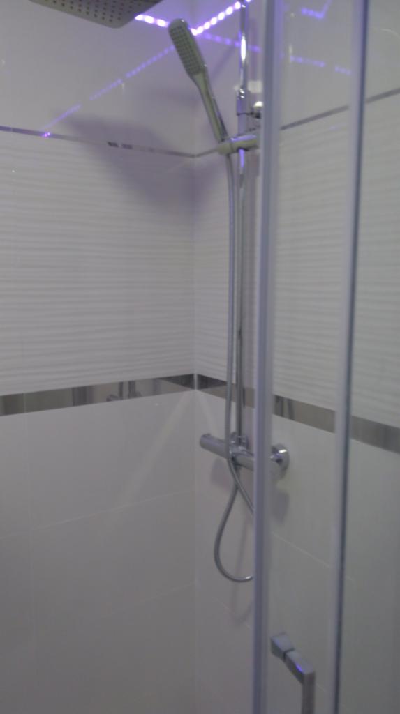 Nowoczesna kabina prysznicowa z deszczownicą i słuchawką, białe płytki z poziomym wzorem i srebrnym paskiem, oświetlenie LED w kolorze fioletowym u góry.