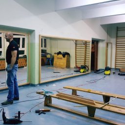 NORDICA - WOJCIECH GROSS - Montaż dużych luster w drewnianych ramach na ścianie sali gimnastycznej. Widoczne narzędzia i ekipa montażowa podczas pracy. Sala z drabinkami gimnastycznymi w tle.