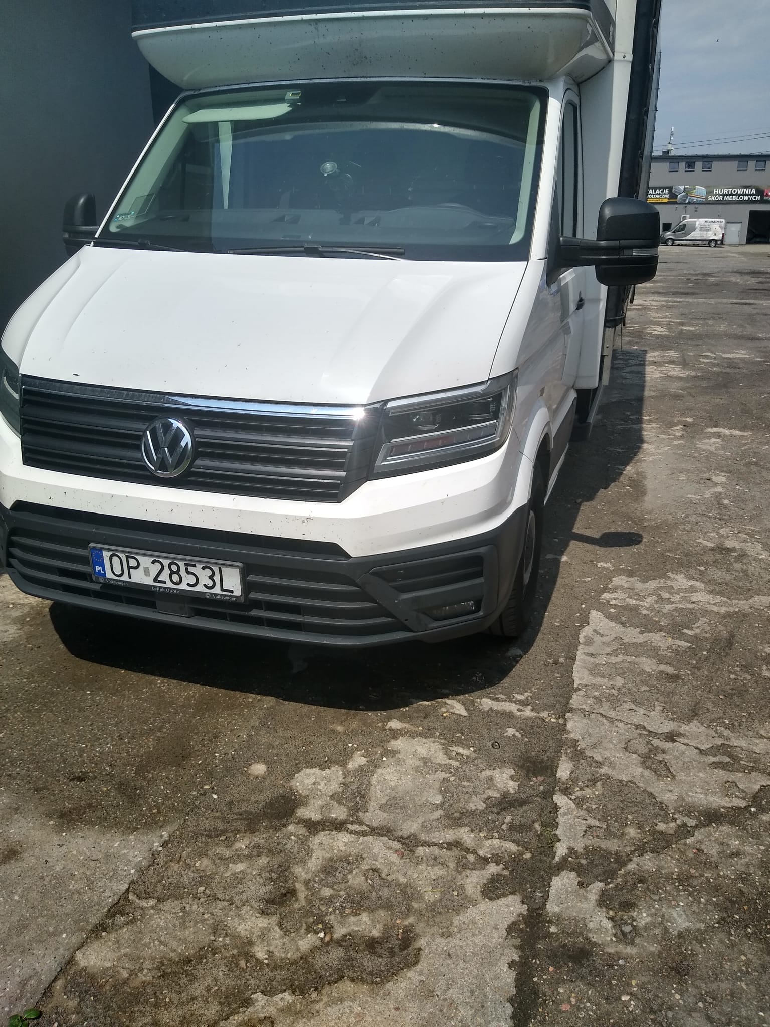 Biały samochód dostawczy Volkswagen Crafter z zabudową kontenerową, zaparkowany na betonowym placu. Widoczna tablica rejestracyjna OP 2853L.