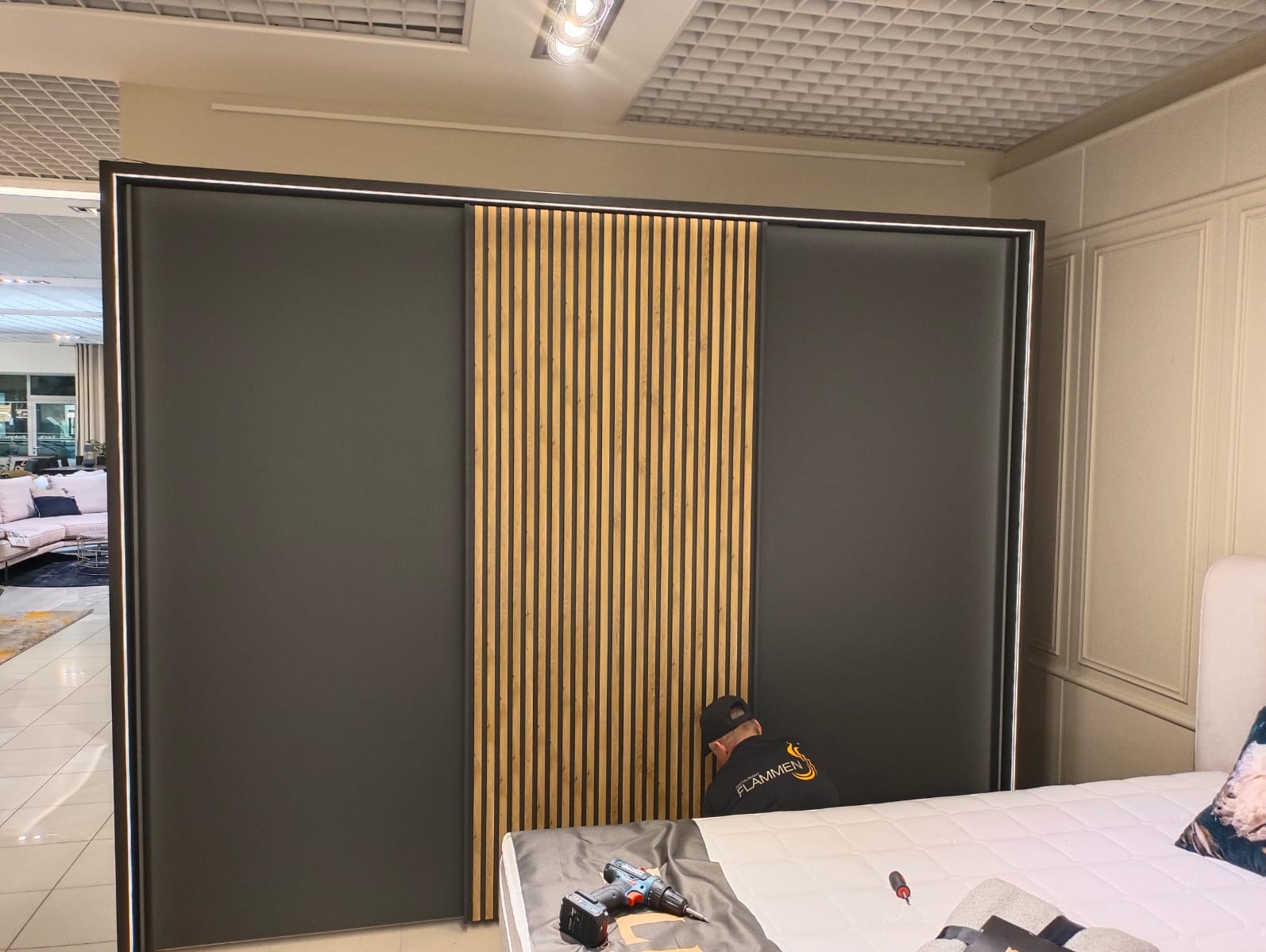 Montaż przesuwnych drzwi szafy z drewnianymi lamelami przez montera w czarnej koszulce z logo na tle jasnej sypialni. Widoczne narzędzia na łóżku.