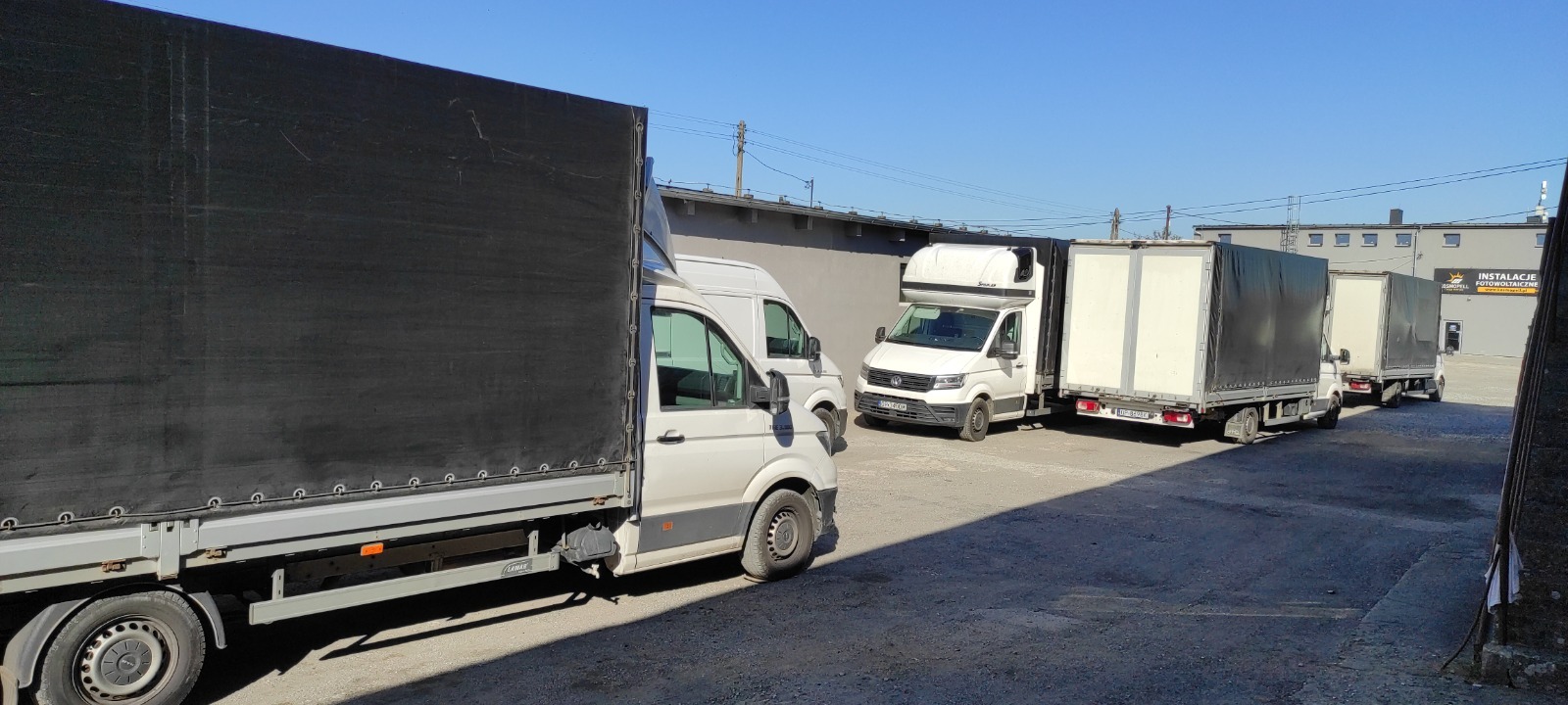 Flota białych busów dostawczych z plandekami i zabudowami, zaparkowana na placu firmy transportowej w słoneczny dzień. Widoczne detale konstrukcyjne pojazdów.