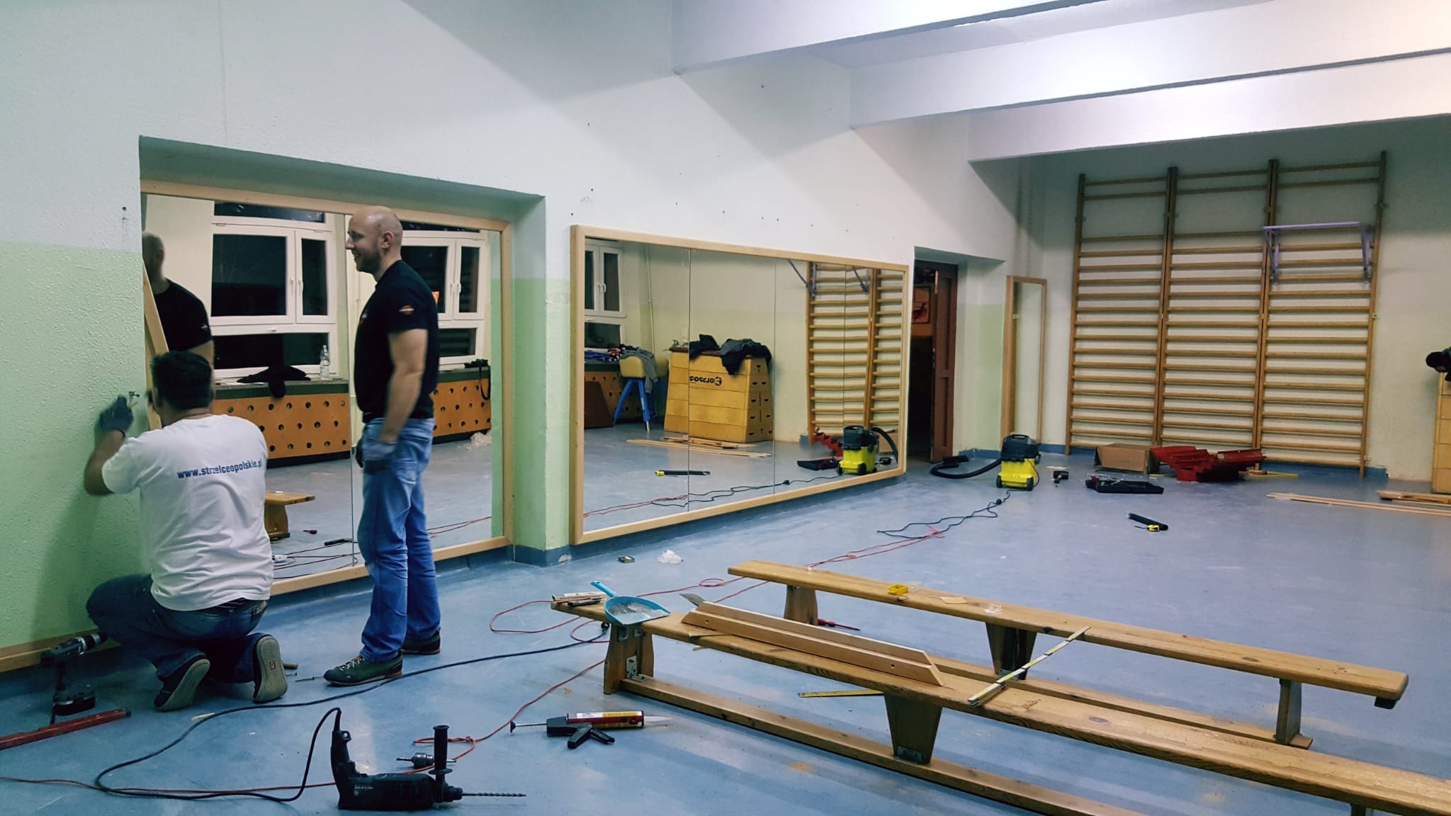 Montaż dużych luster w drewnianych ramach na ścianie sali gimnastycznej. Widoczne narzędzia i ekipa montażowa podczas pracy. Sala z drabinkami gimnastycznymi w tle.