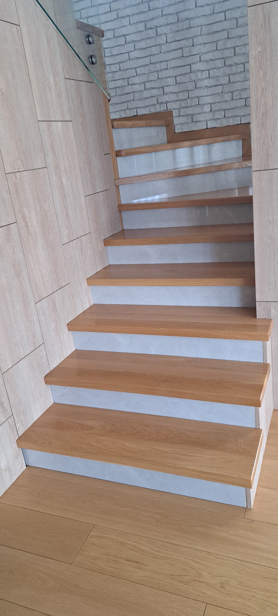 Drewniane schody z jasnymi podstopnicami i szklaną balustradą, widok z dołu, z fragmentem drewnianej podłogi i ściany z imitacji cegły w tle.