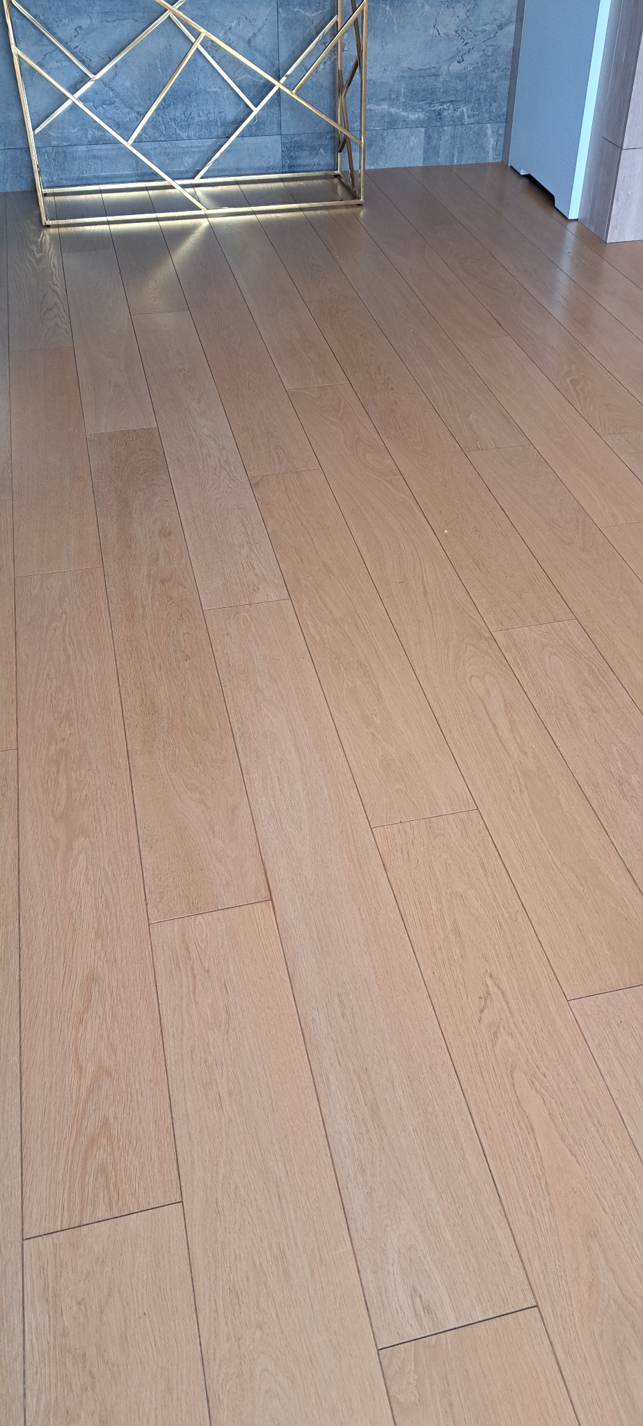 Jasny parkiet ułożony w regularny wzór, widoczny fragment złotej konsoli i szara ściana w tle. Podłoga po renowacji, z widoczną strukturą drewna i delikatnym połyskiem.