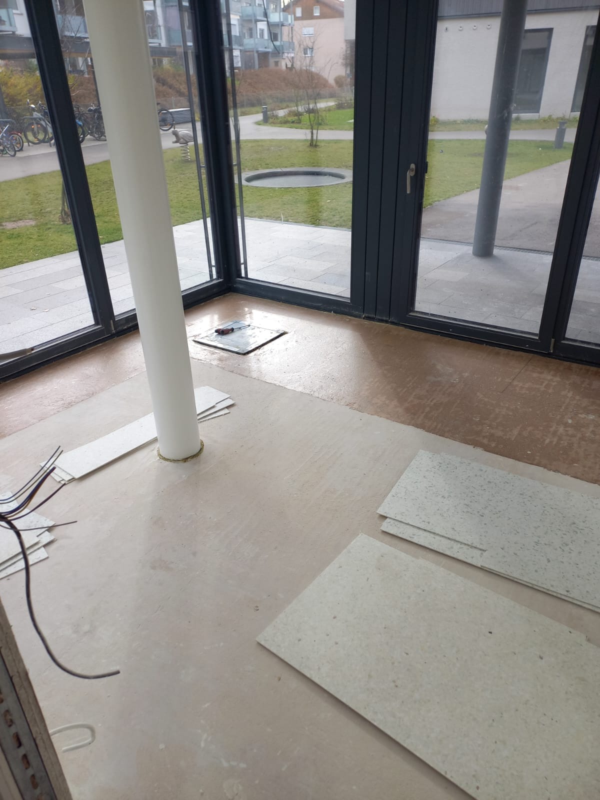 Przygotowanie do ułożenia jasnych paneli podłogowych w pomieszczeniu z dużymi oknami i kolumną. Częściowo widoczna stara podłoga i narzędzia.