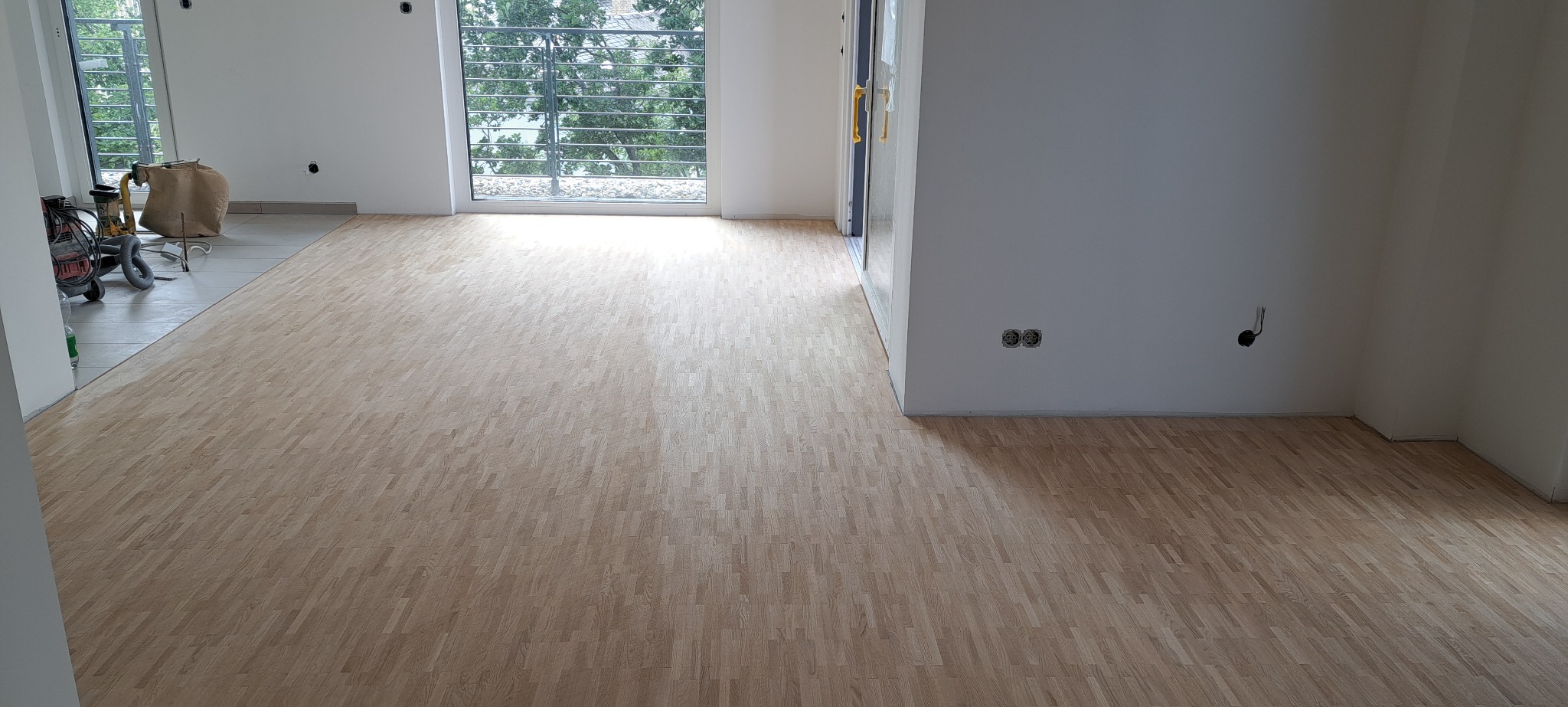 Jasny parkiet ułożony w jodełkę w przestronnym, jasnym pokoju z dużym oknem. W tle widoczny sprzęt do cyklinowania i worek z pyłem.