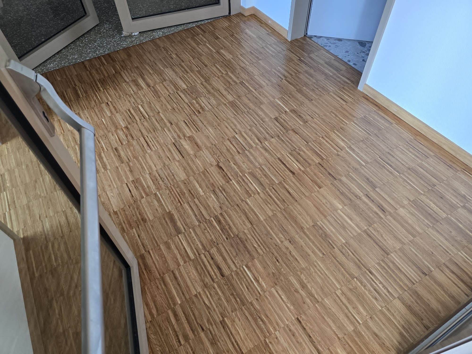 Parkiet ułożony w jodełkę, widok z góry, z fragmentem otwartych drzwi po lewej stronie. Jasne drewno, regularny wzór, świeżo po renowacji, z połyskiem.