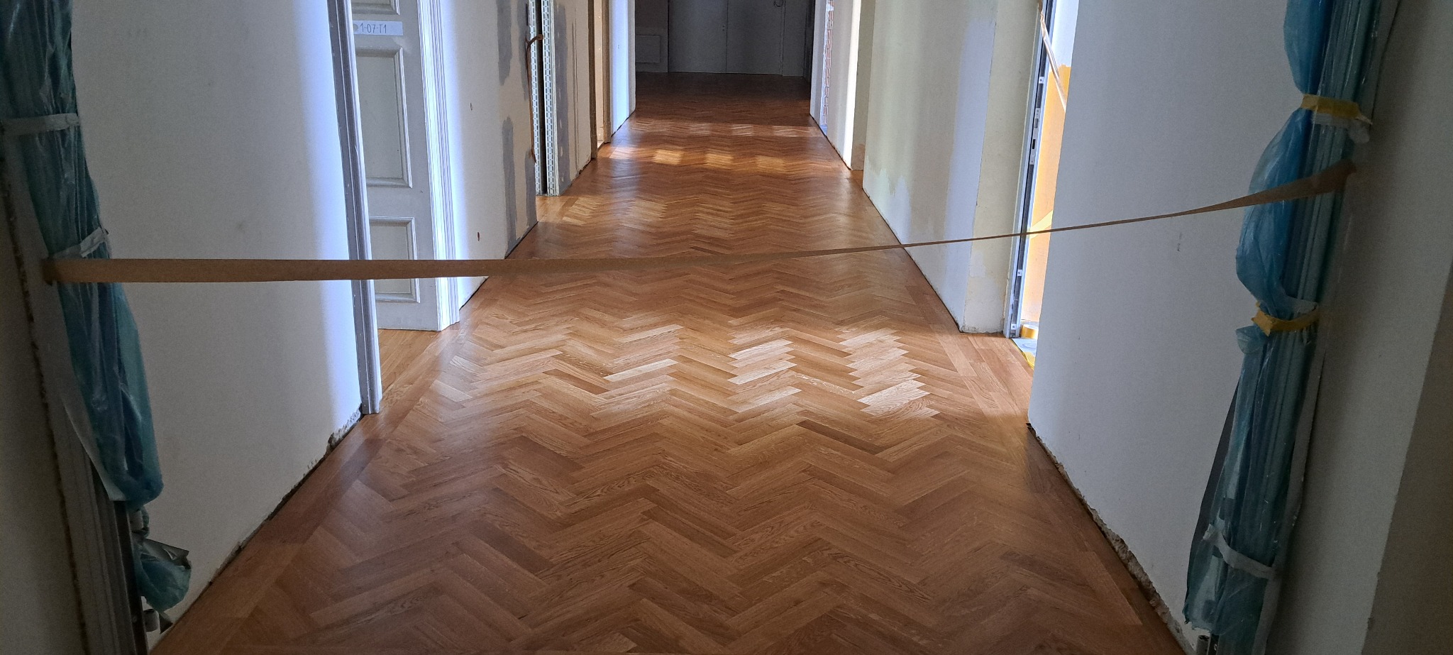 Długi korytarz z parkietem ułożonym w jodełkę, zabezpieczony taśmą. Widoczne odnowione podłogi w jasnym odcieniu drewna, ściany przygotowane do malowania.