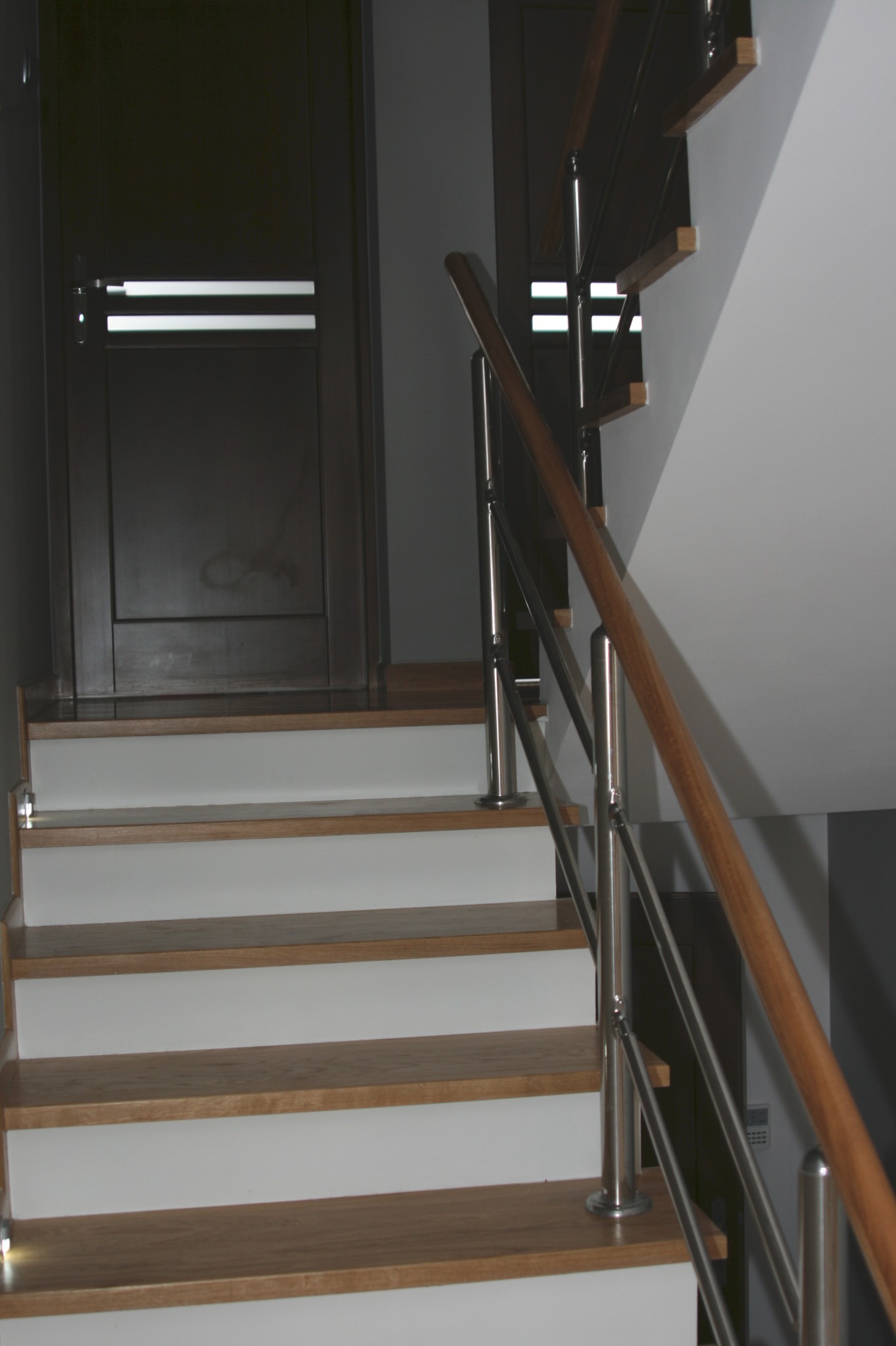 Nowoczesne schody drewniane z białymi podstopnicami i metalową balustradą, oświetlone punktowo, prowadzące do ciemnych drzwi z poziomymi przeszkleniami.