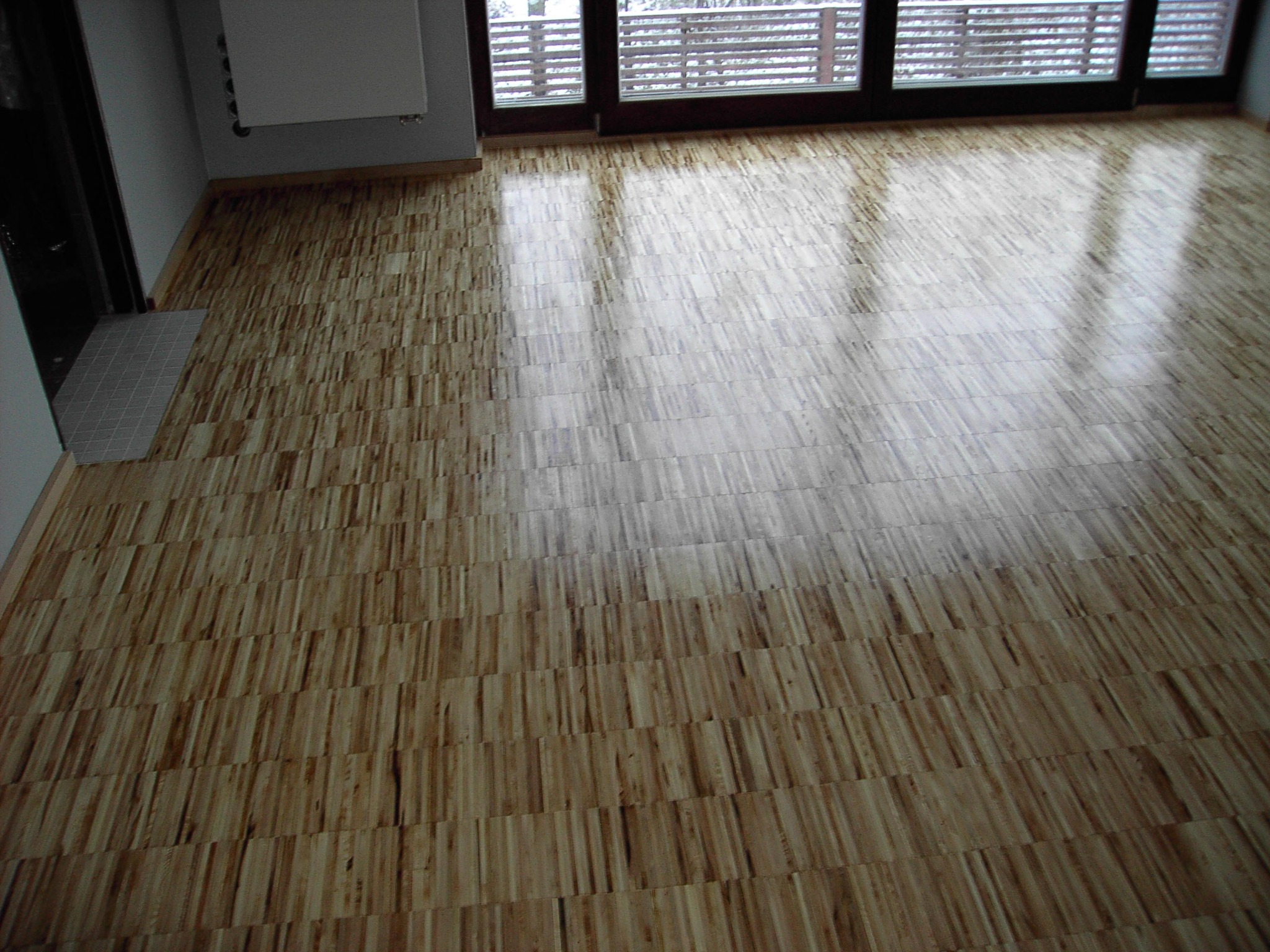 Jasny parkiet ułożony w regularny wzór, odbijający światło z okna balkonowego. Widoczny fragment grzejnika i drzwi. Podłoga po renowacji.