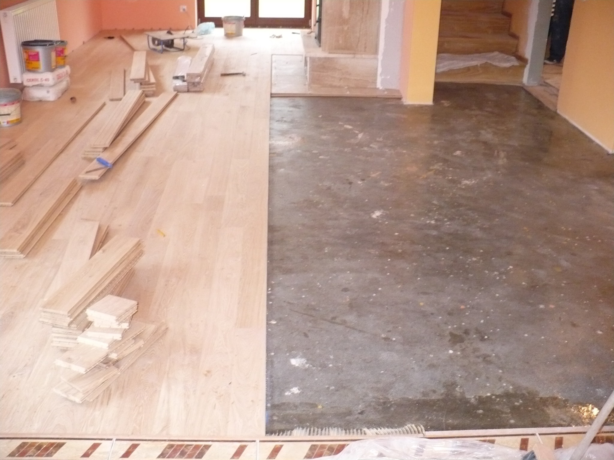 Połowa podłogi z surowym betonem, druga z jasnym parkietem w trakcie układania. Widoczne deski, narzędzia i materiały budowlane. Ujęcie z góry, w tle grzejnik.