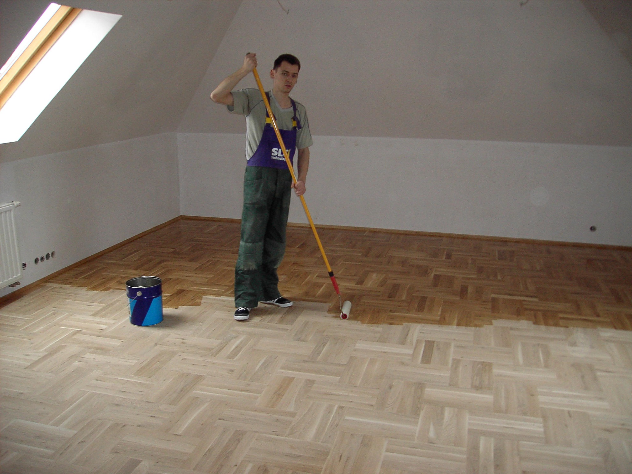 Parkiet w jodełkę, częściowo pokryty lakierem. Mężczyzna z wałkiem maluje podłogę na poddaszu. Widoczne okno dachowe i wiadro z farbą.