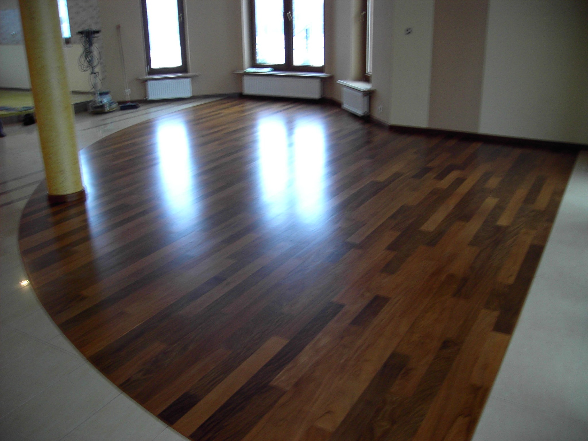 Parkiet z ciemnego drewna ułożony w jodełkę, z widocznym połyskiem i fragmentem kolumny w tle. Podłoga łączy się z jasną, ceramiczną posadzką.