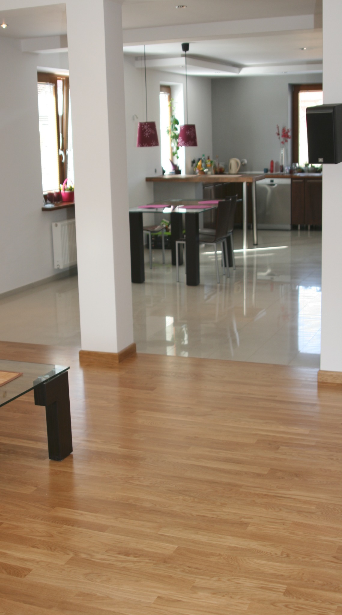 Jasny parkiet w salonie z widokiem na kuchnię. Podłoga po cyklinowaniu, gładka powierzchnia. Fragment szklanego stolika kawowego. Nowoczesne wnętrze.